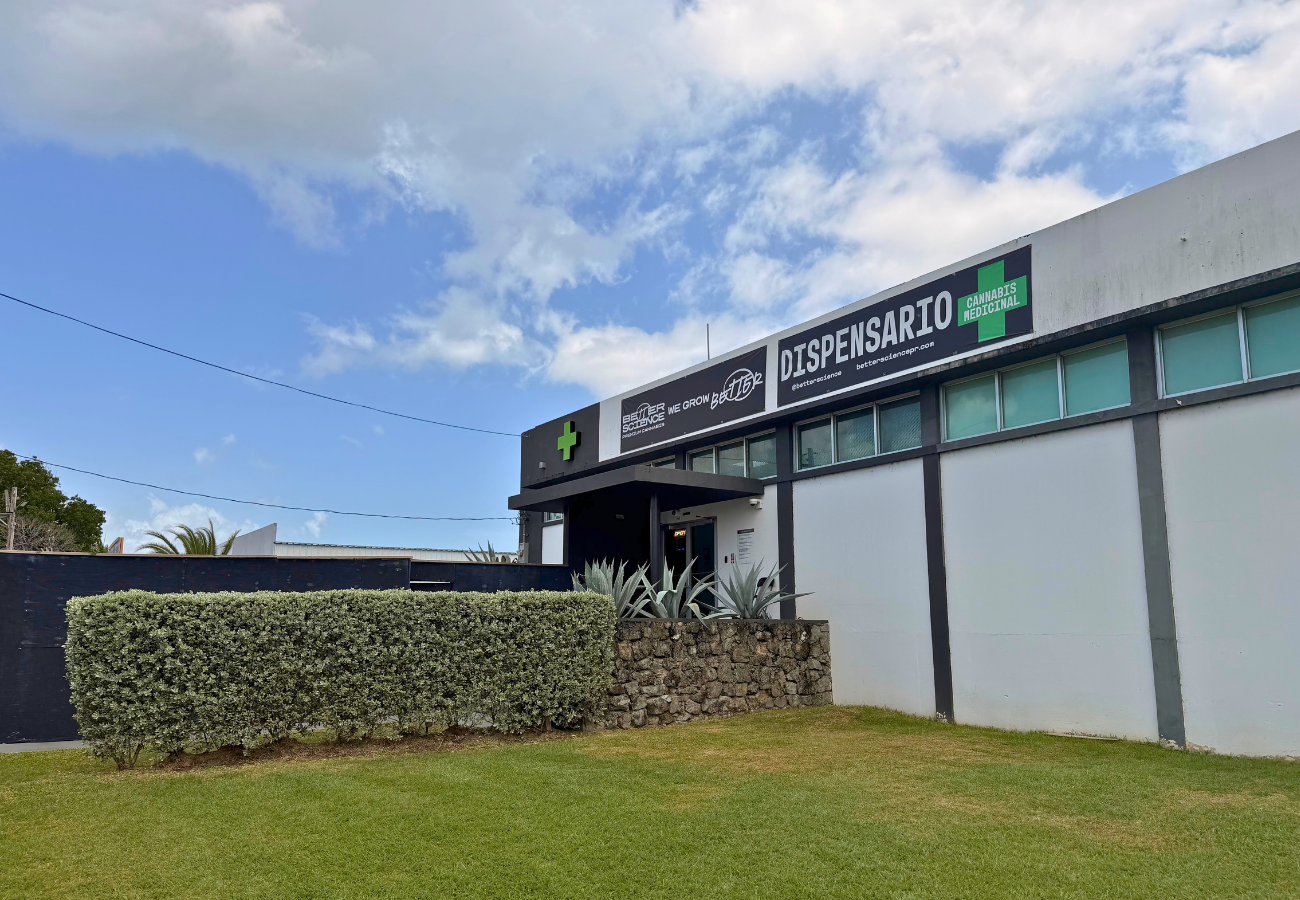 Fachada de una dispensario de cannabis medicinal con señalización en inglés y verde, césped y arbustos en primer plano.