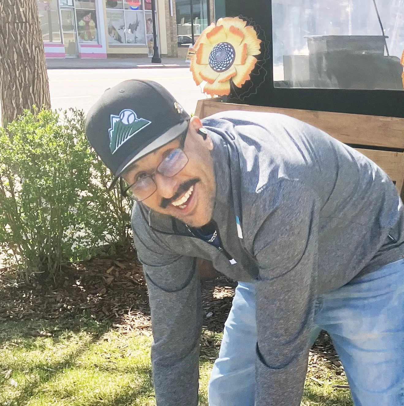 A man wearing glasses, a black cap with a mountain and baseball logo, and a gray jacket, smiling and bending forward outdoors near bushes with a storefront and decorative sunflower cutouts in the background.