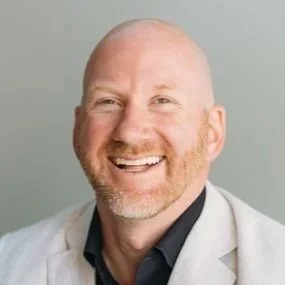 Patrick Weeg headshot (A smiling man, wearing a light-colored blazer and a dark shirt, against a plain background.)