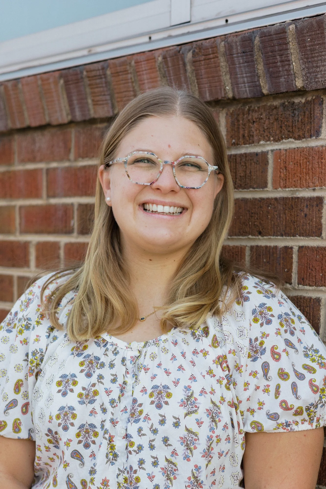 Sarah Margaret outside smiling while standing in front of a red brick wall, wearing glasses and a white floral printed shirt