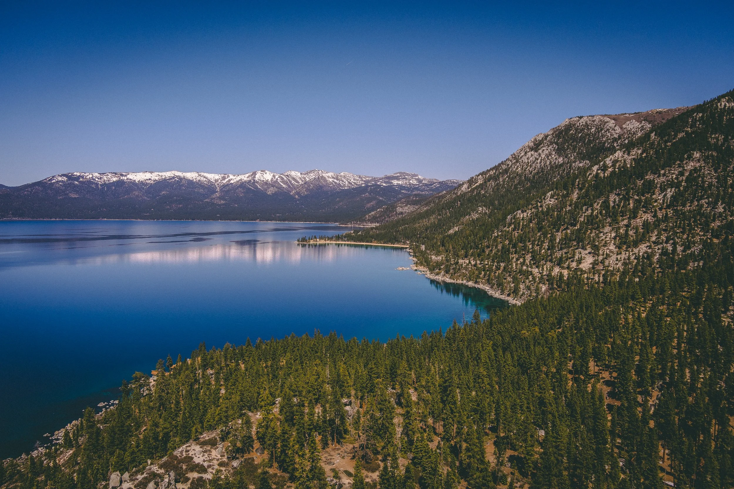 Aerial view of Lake Tahoe with surrounding pine forests and distant snow-capped mountains under a clear blue sky.