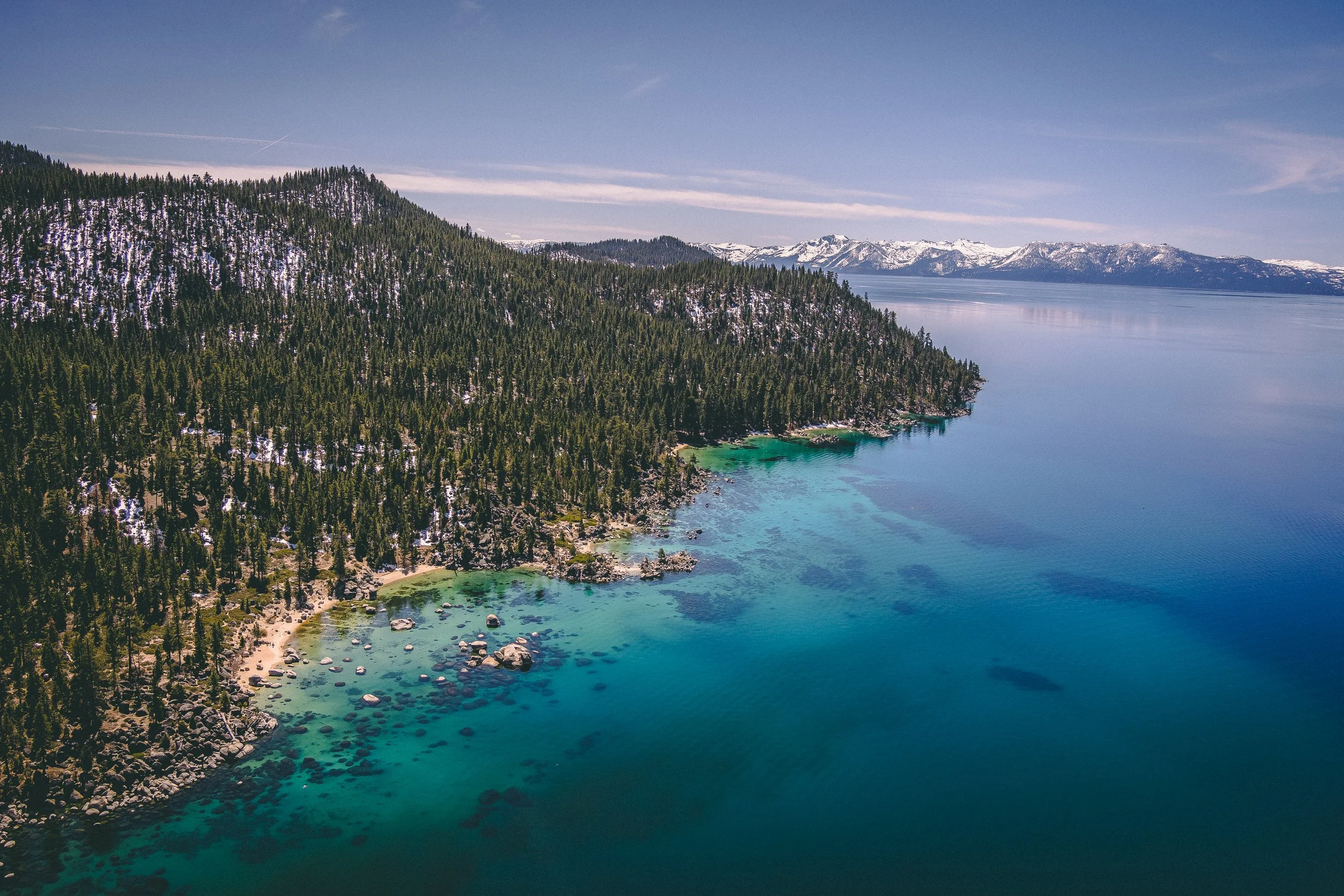 Aerial view of a forested mountain landscape beside a clear blue lake with a sandy shoreline and some rocks.