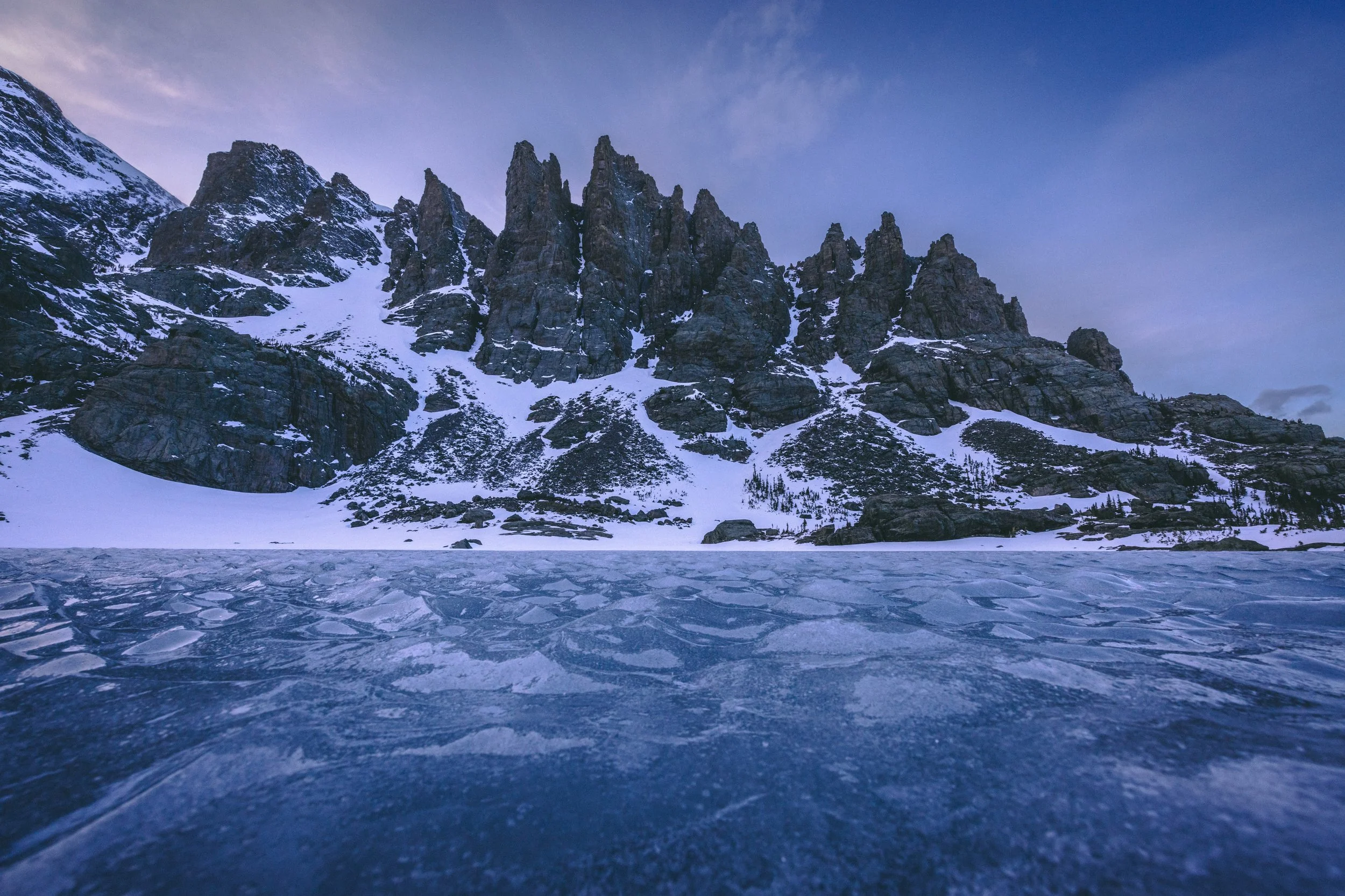 Rocky mountain peaks covered in snow with a frozen lake in the foreground under a cloudy sky.