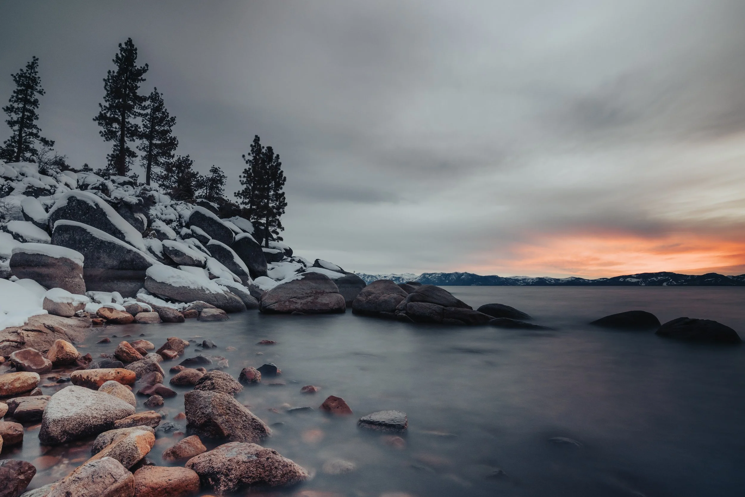 Snow-covered rocks and trees by a calm lake at sunset, with mountains in the distance and a cloudy sky.
