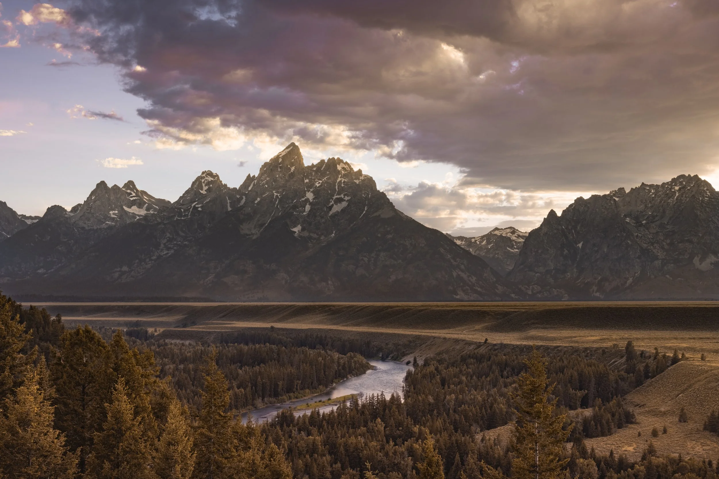 Scenic view of the Grand Teton mountain range at sunrise with a river and forest in the foreground.