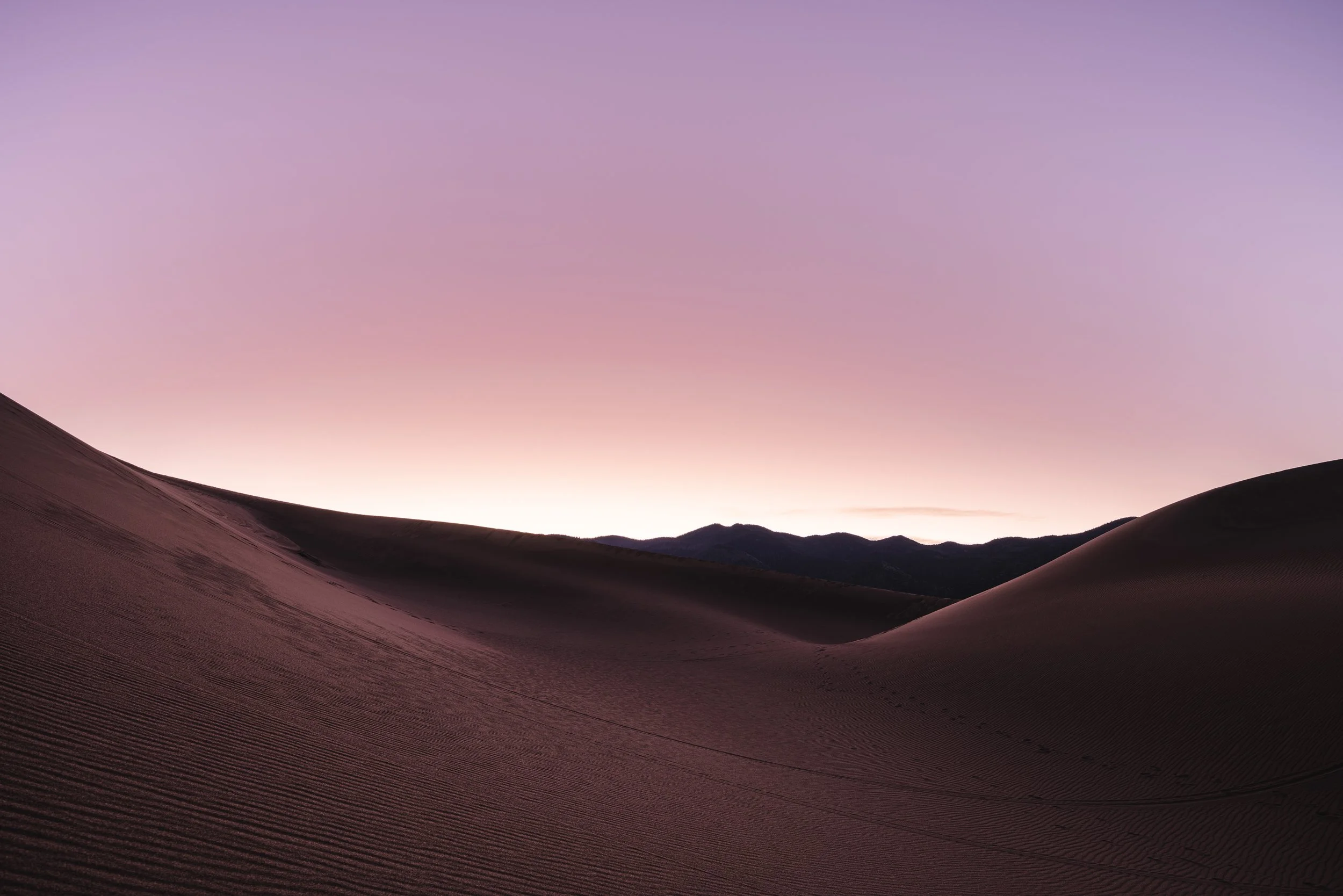 Sand dunes at sunset with a purple sky and distant mountains.