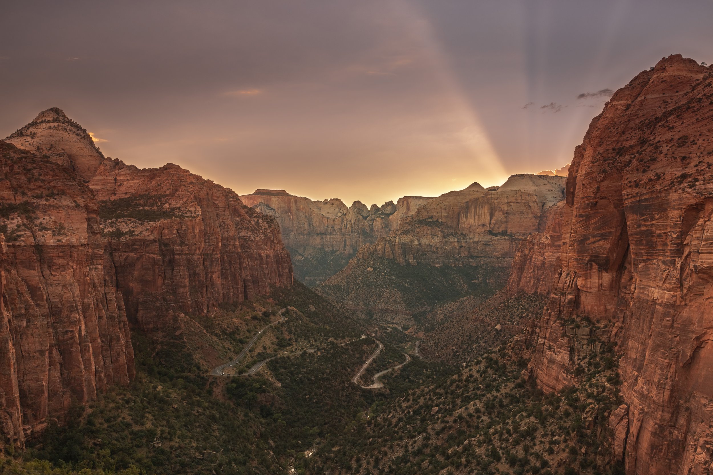 Scenic view of Zion Canyon at sunset in Zion National Park, featuring rugged red cliffs and a winding road through the valley.