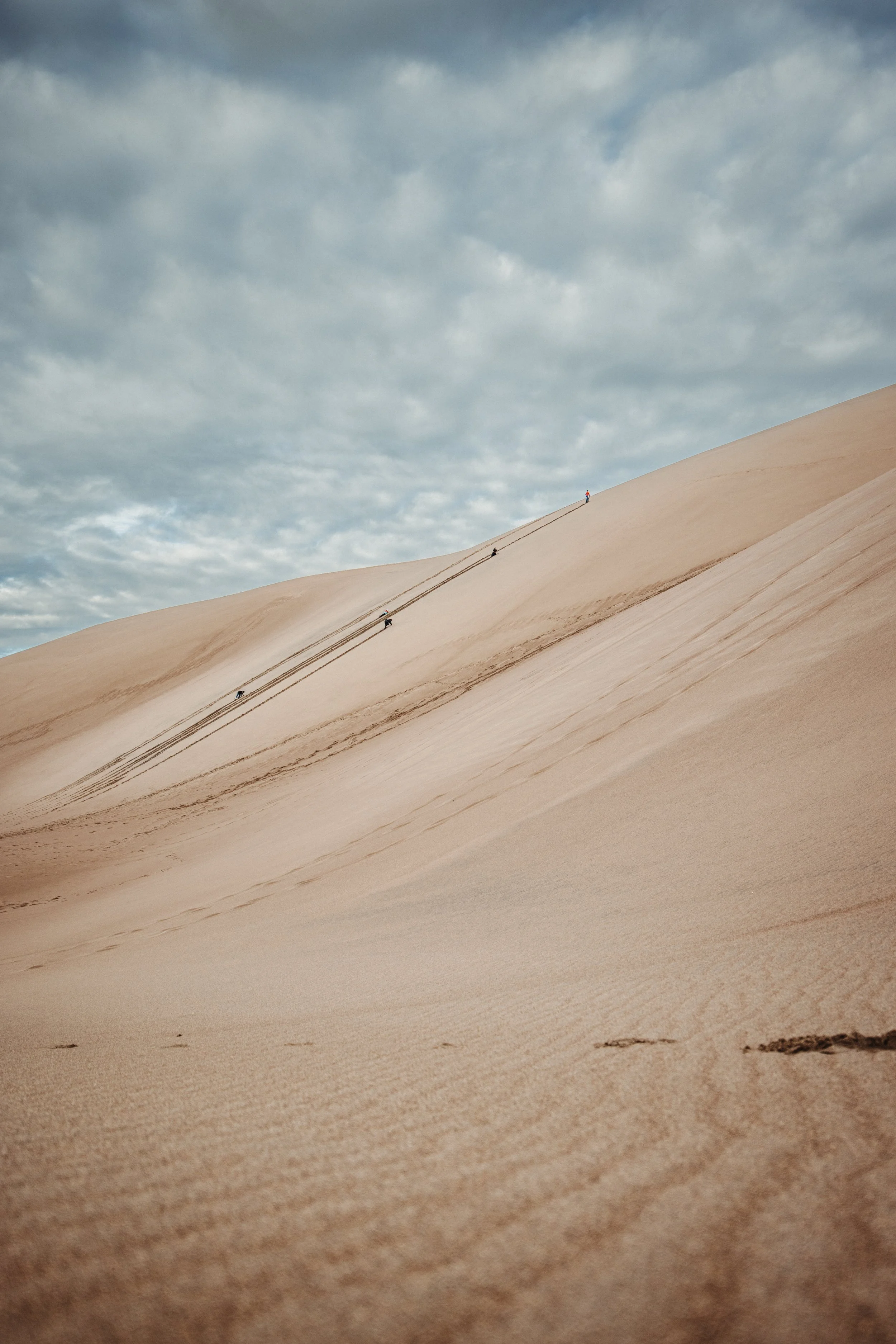 People climbing a large sand dune under a cloudy sky.