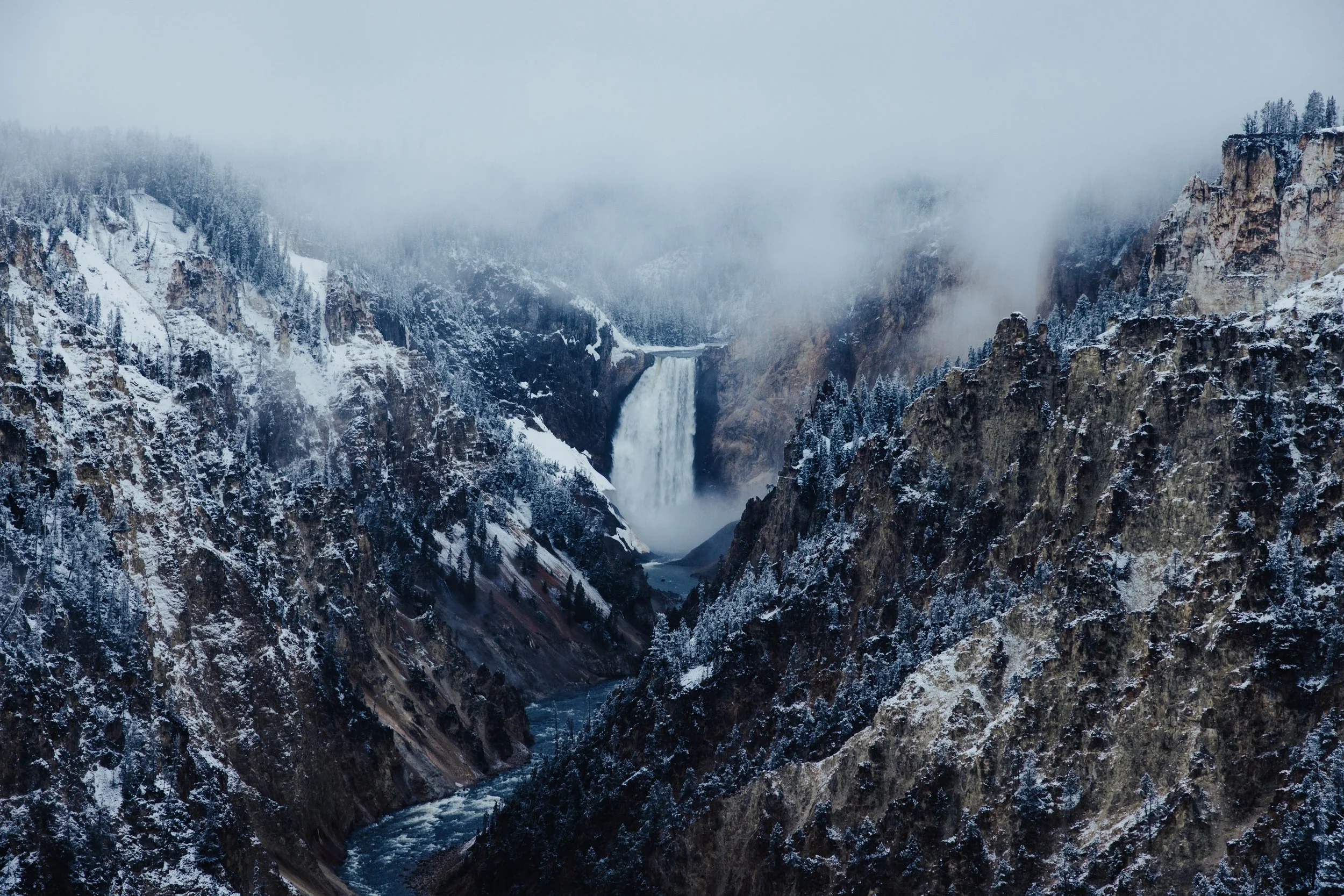 Snow-covered canyon with a waterfall cascading down, surrounded by misty pine trees.