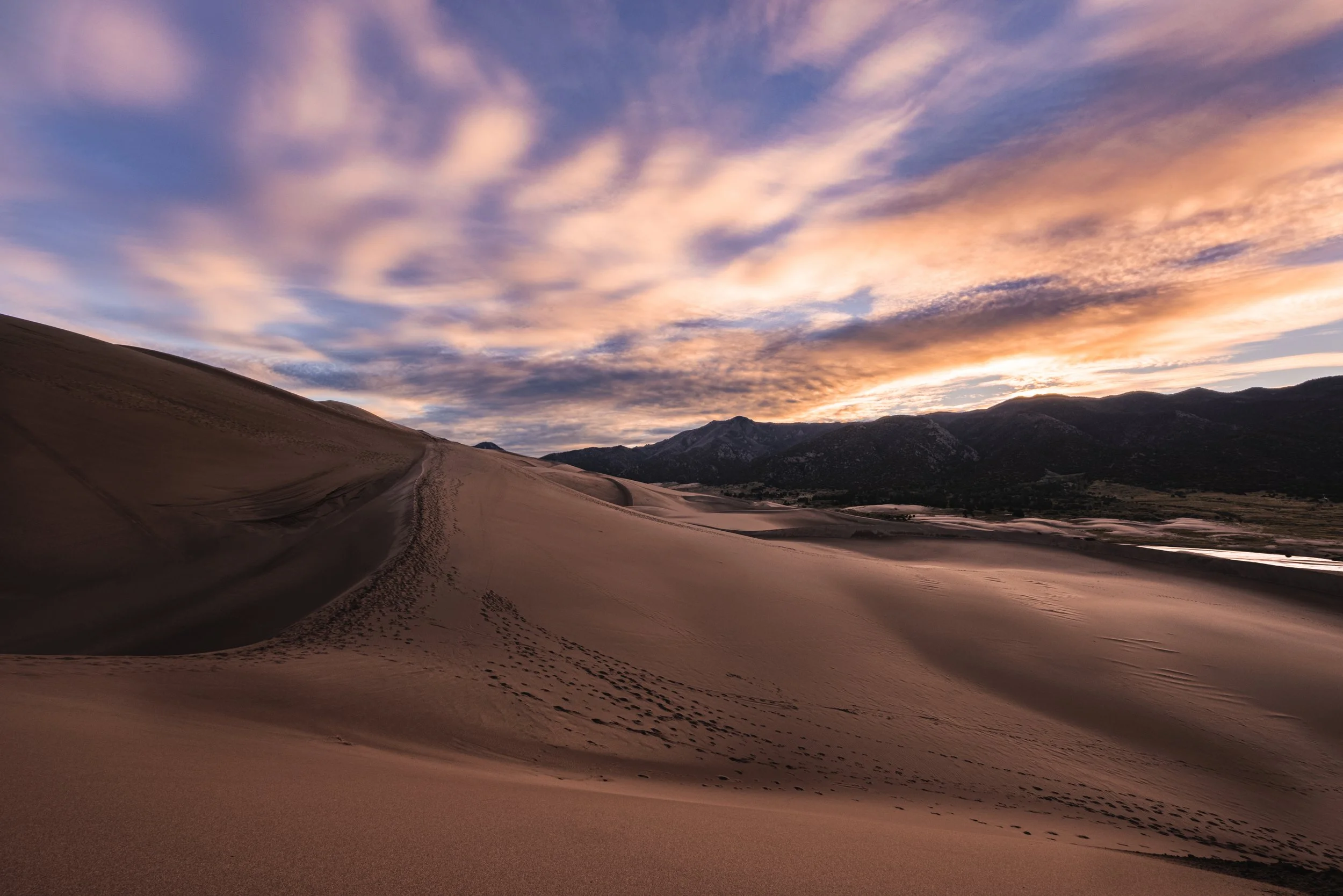 Sand dunes at sunset with mountains in the background and a colorful sky overhead.