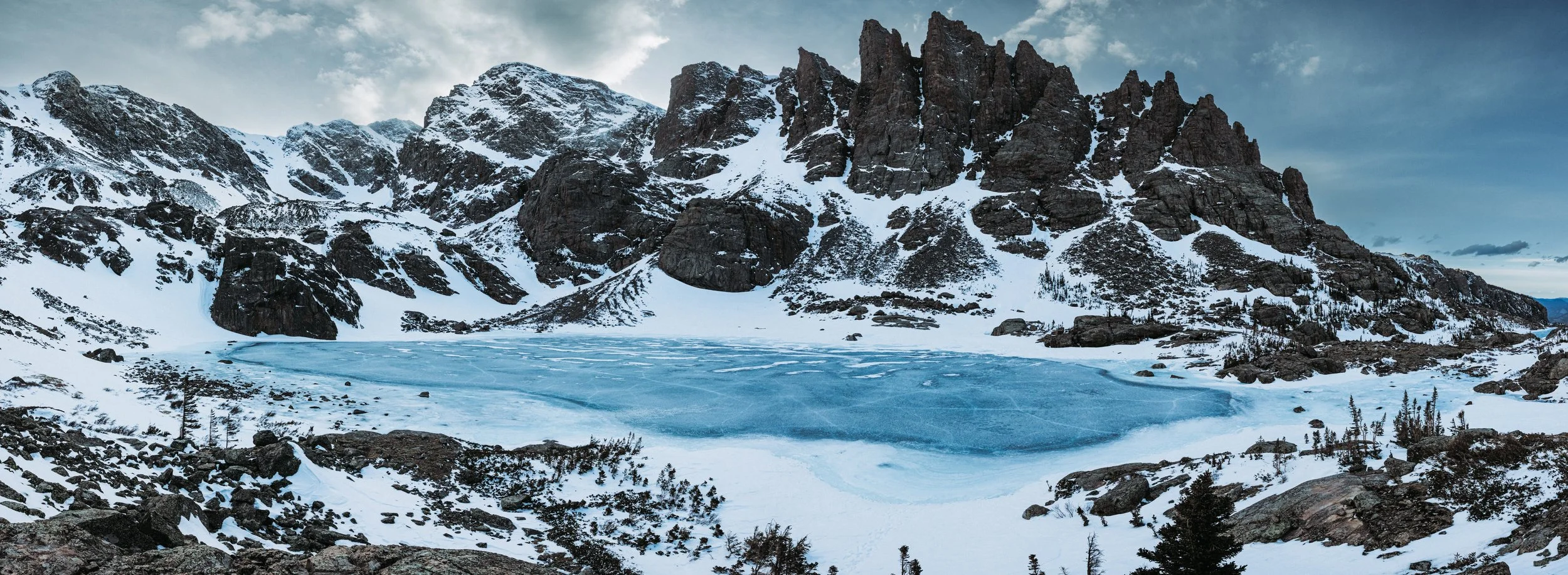 Frozen alpine lake surrounded by snow-covered mountains and rocky peaks, with a cloudy sky above.