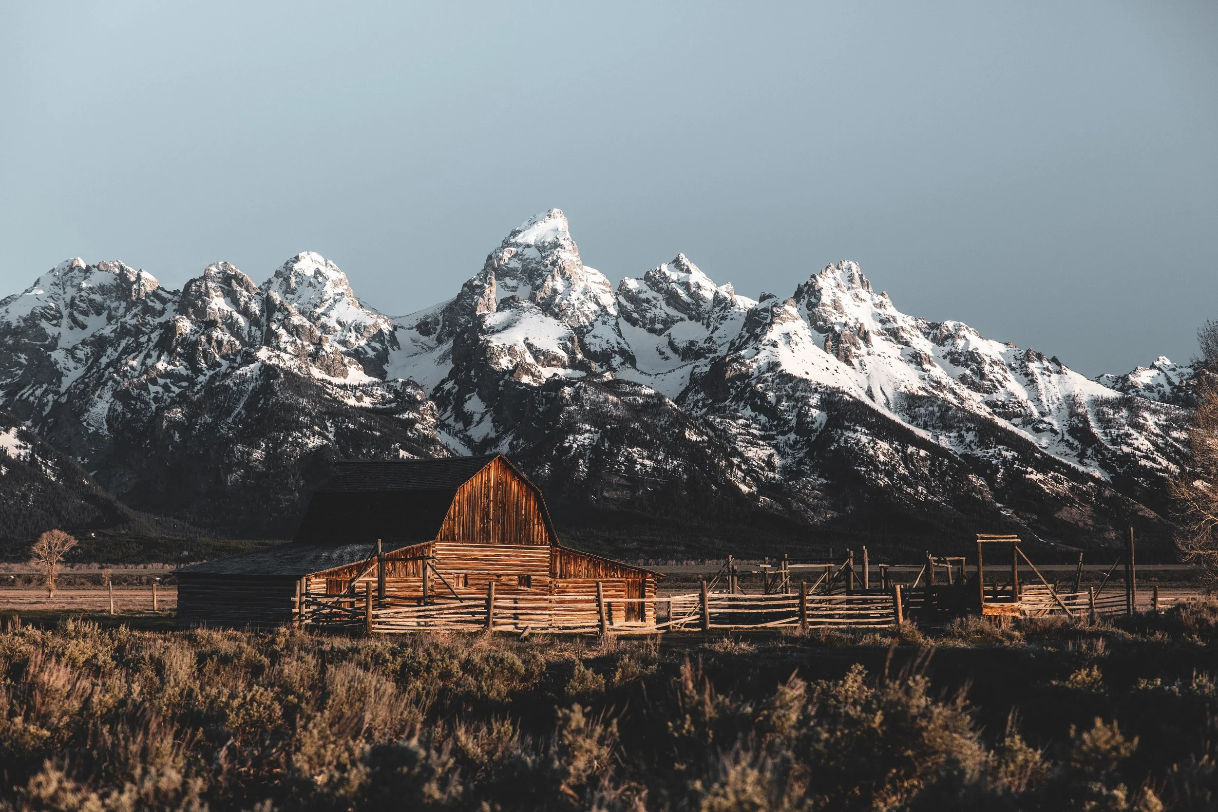 Wooden barn in front of snow-covered mountains under a clear sky.
