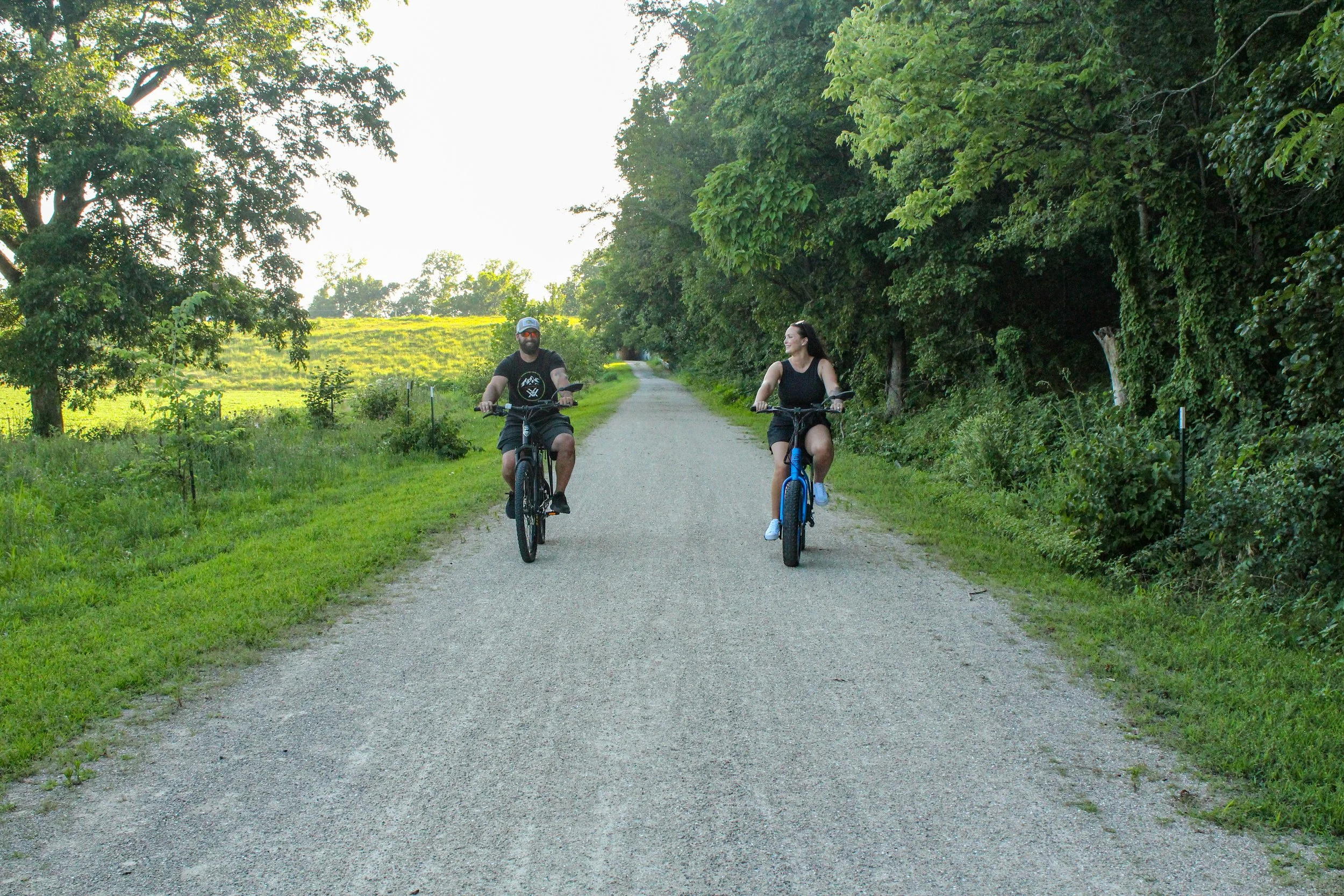Two people riding bicycles on a gravel path surrounded by green trees and grass, with sunlight shining through the foliage.