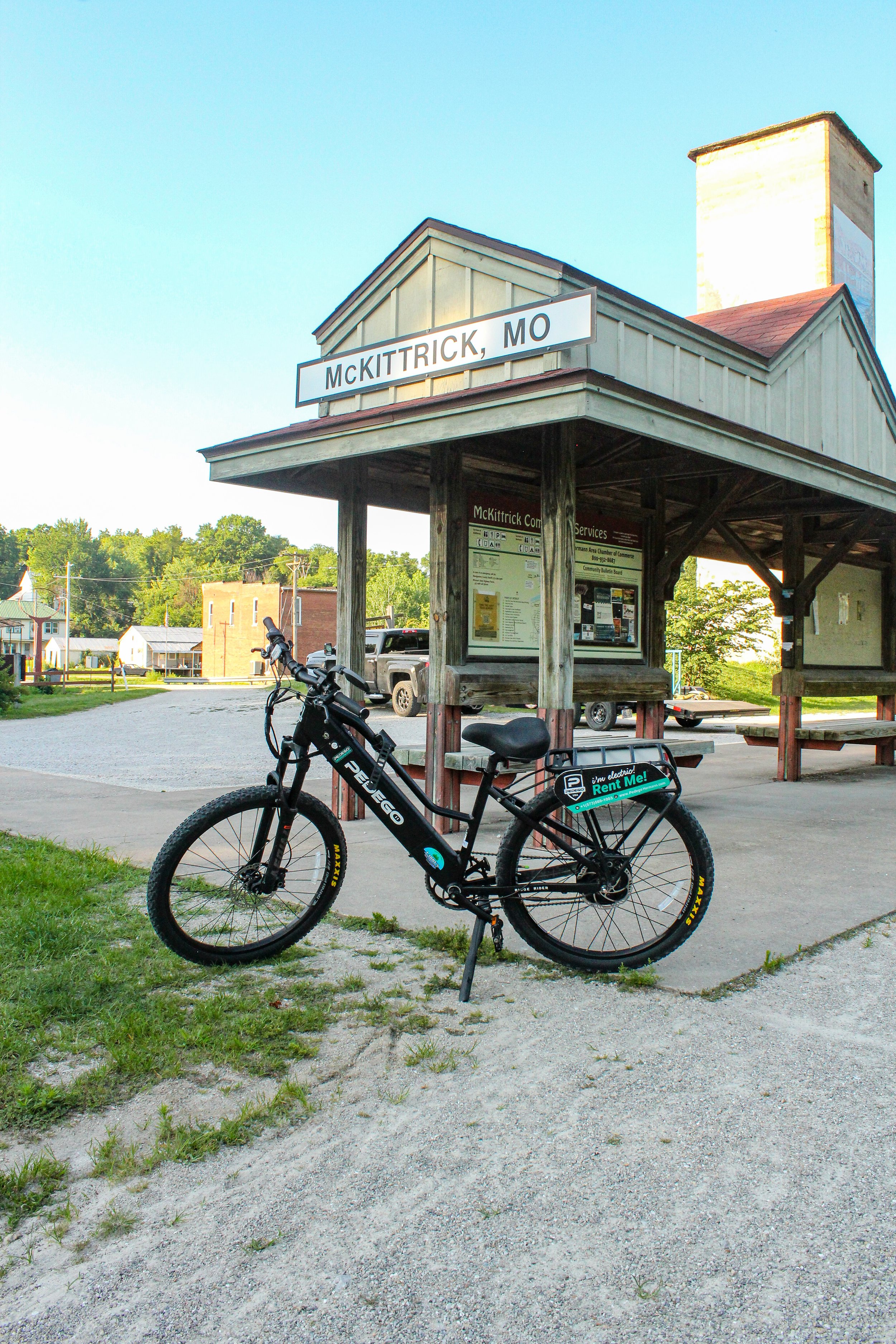 A black electric mountain bike parked in front of a wooden pavilion in McKittrick, Missouri, with a blue sky and trees in the background.