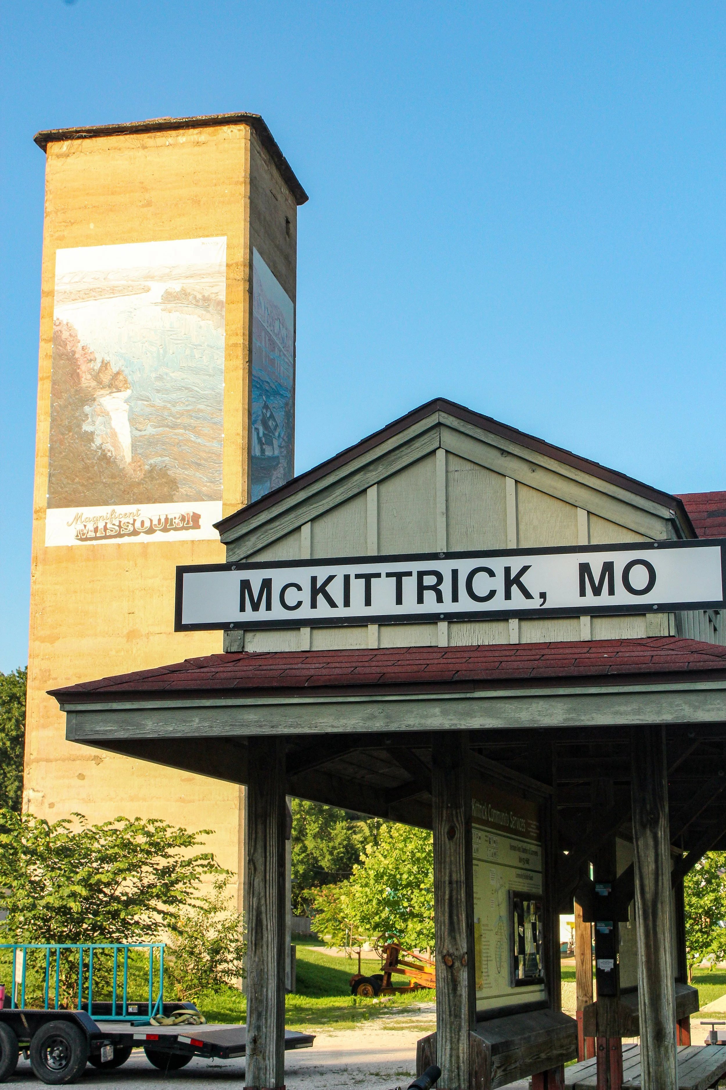 A small wooden shelter with a corrugated roof and a sign reading 'McKitttrick, MO'. Behind it, there is a wall with a large vintage poster depicting a scenic landscape and the words 'Magnificent Missouri'. There is a trailer with some equipment and trees and greenery in the background.