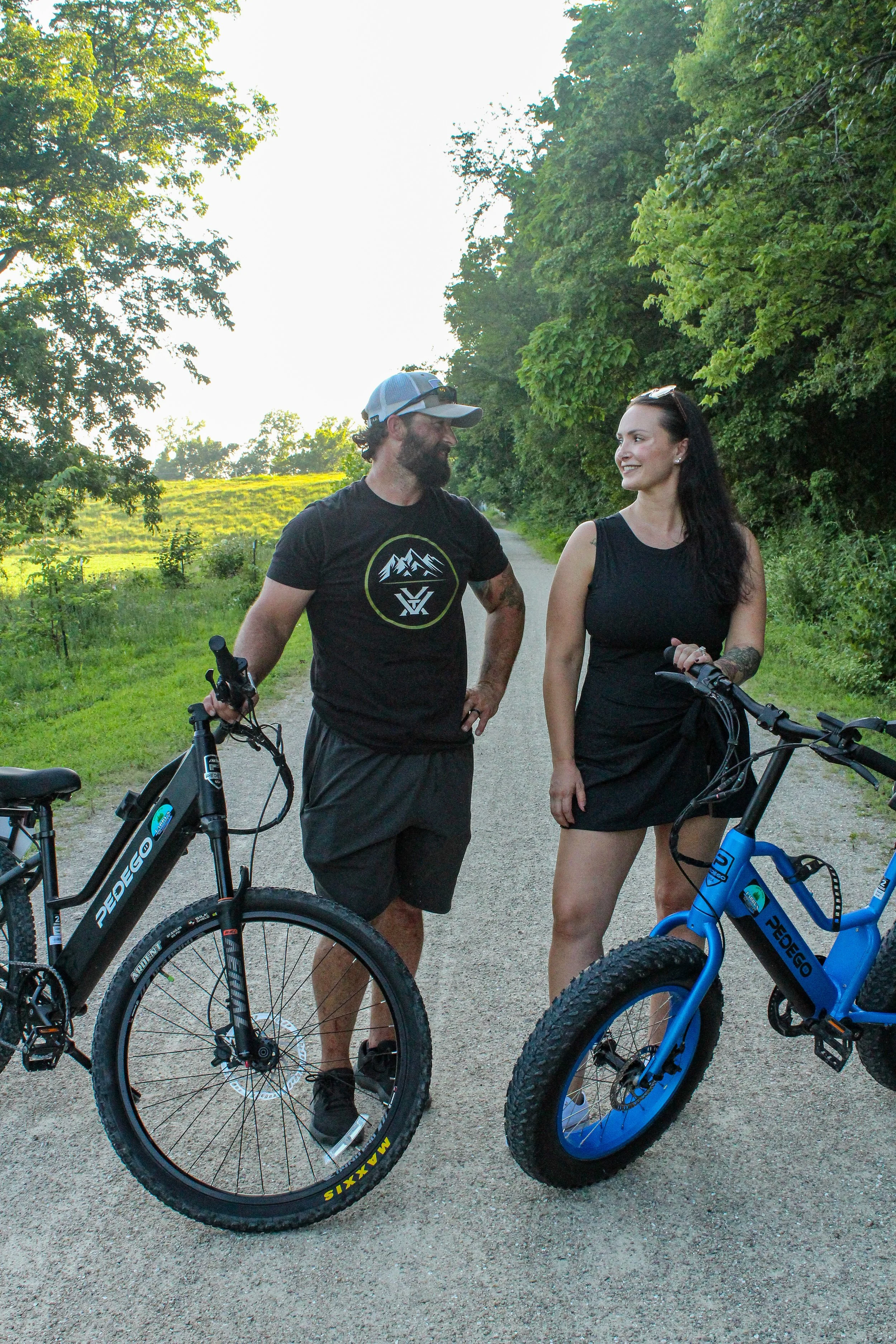 Two people with bicycles standing on a dirt path surrounded by lush green trees under a bright sky, smiling at each other.