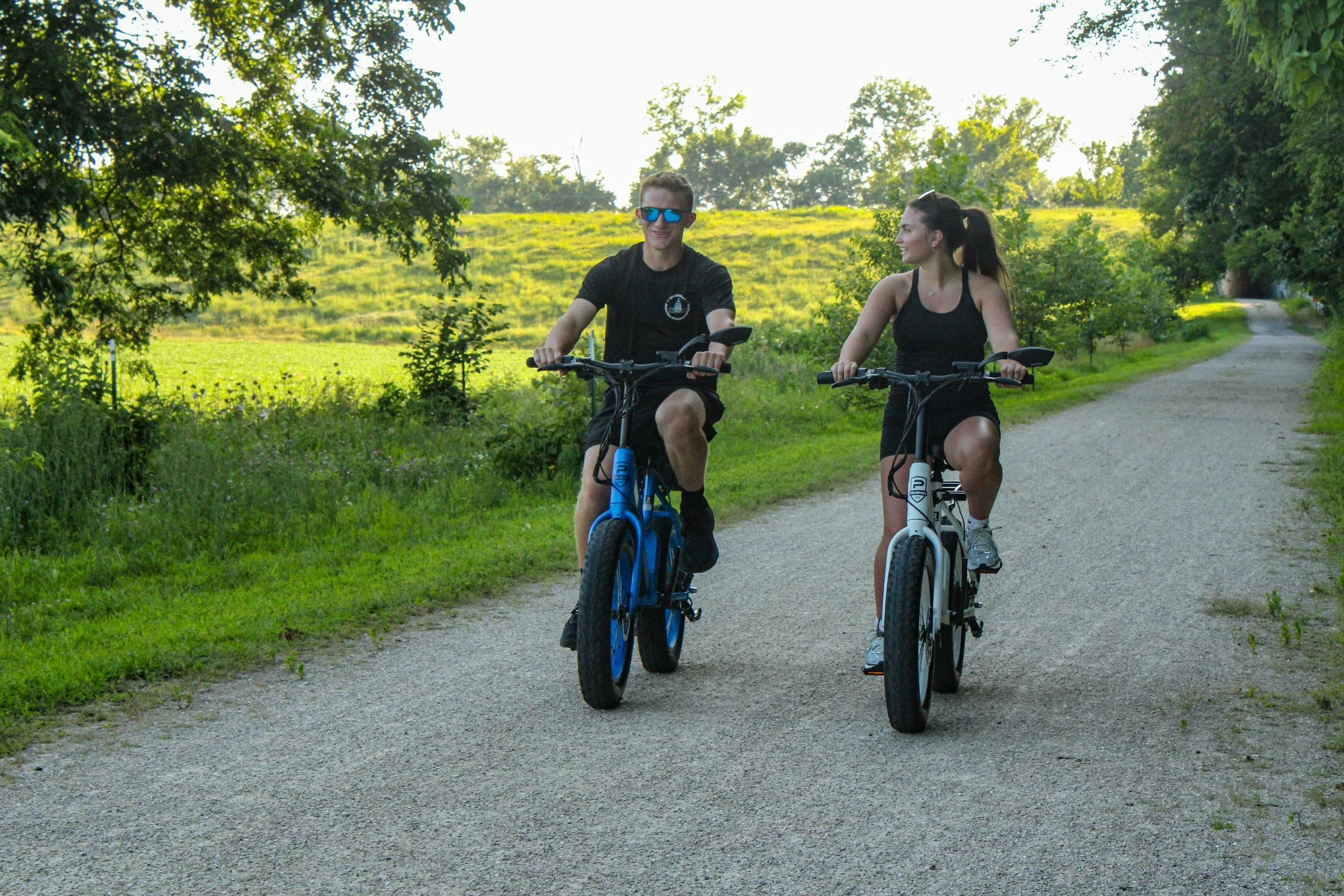 A man and a woman riding bikes on a gravel path through a lush green park with trees and open grassy fields in the background during daytime.