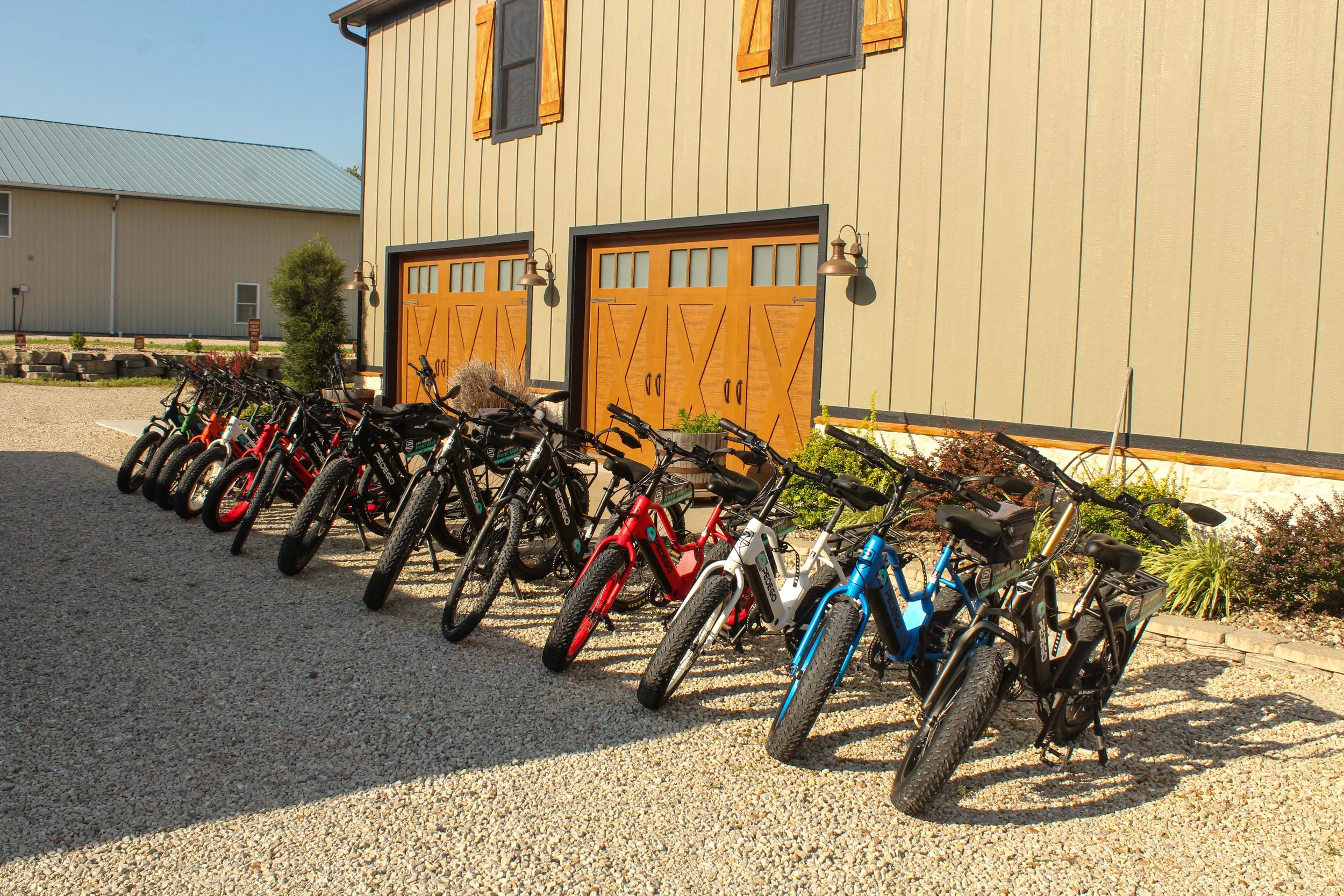 A row of bicycles parked outside a building with beige vertical siding, wooden garage doors, and outdoor wall-mounted lamps.