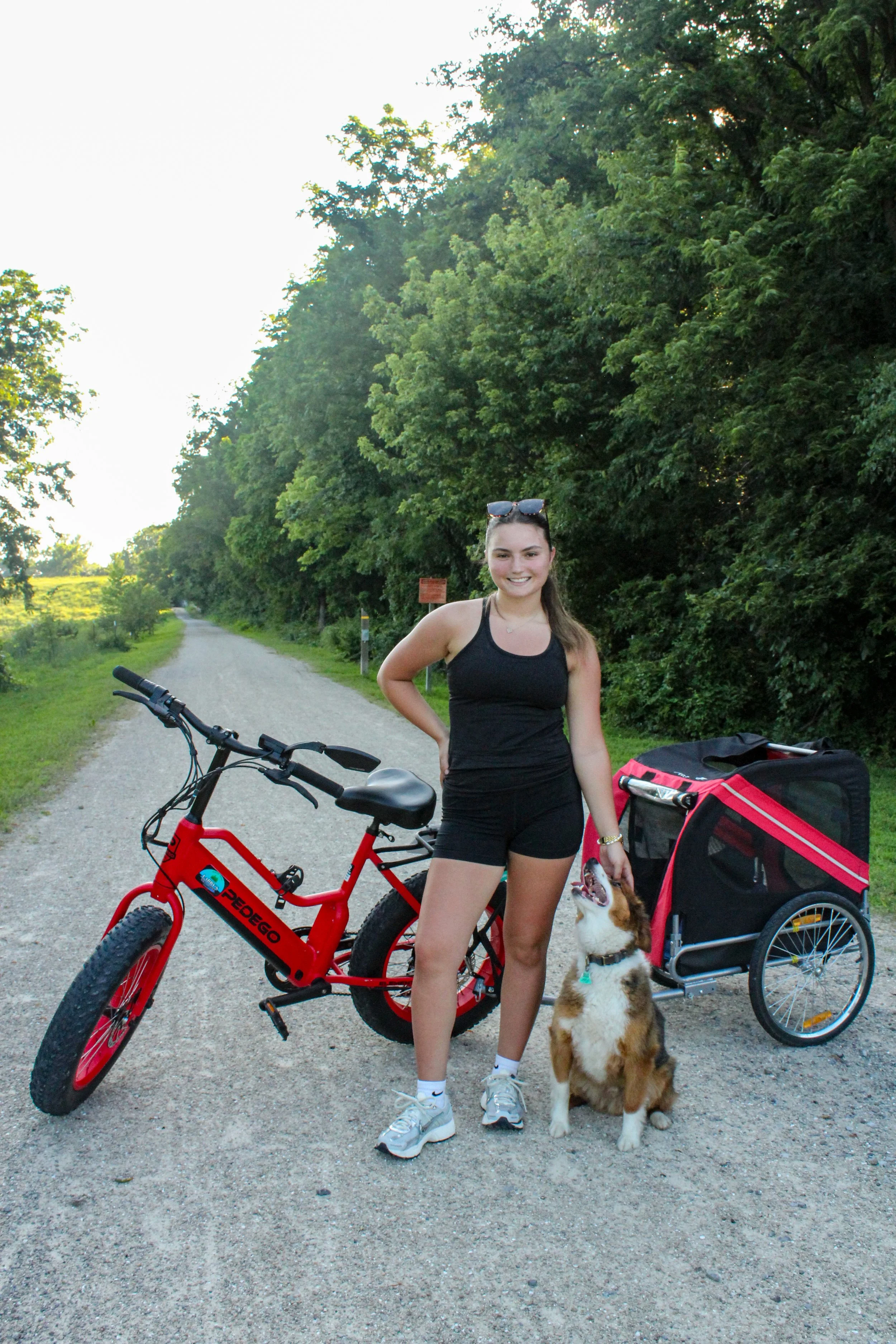 A young woman standing next to a red electric bike on a gravel road, with a dog sitting beside her, in a green, wooded outdoor area during daytime.