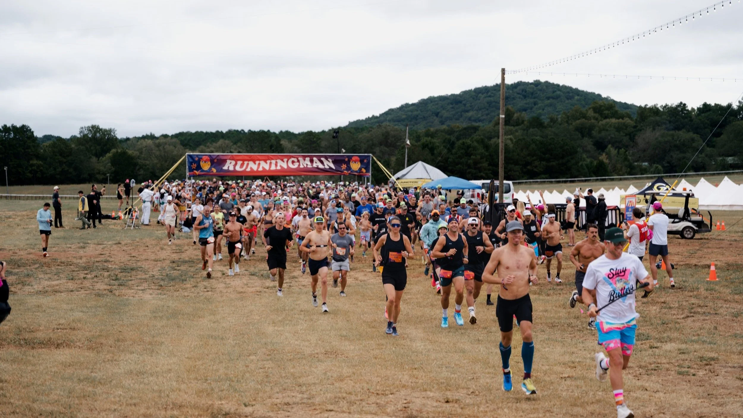 A large group of runners participating in a race, emerging from the starting line marked 'Running Man', on a field with mountains in the background, overcast sky, and tents along the side.