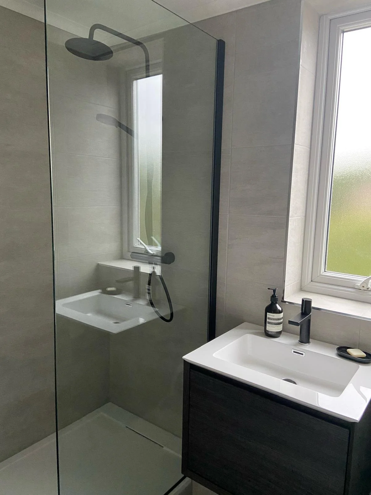 Modern bathroom with a walk-in shower, glass partition, gray tiles, a white sink with a black cabinet, a soap dispenser, and a mirror above the sink. A window provides natural light.