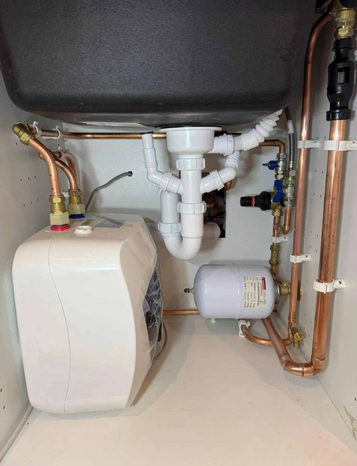 Under-sink plumbing with white PVC pipes, copper pipes, a gray sink, a water filter, and a small water pressure tank in a laundry or utility closet.