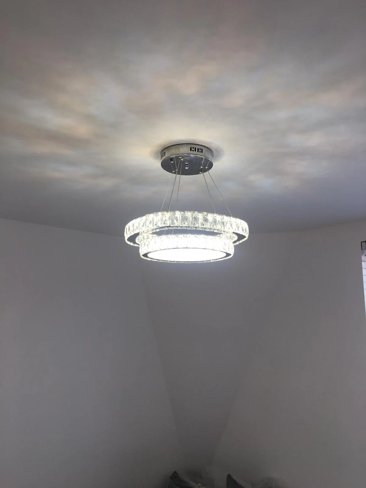 A modern ceiling chandelier with two circular rings of crystal-like material hanging from a ceiling installation with embedded lighting.