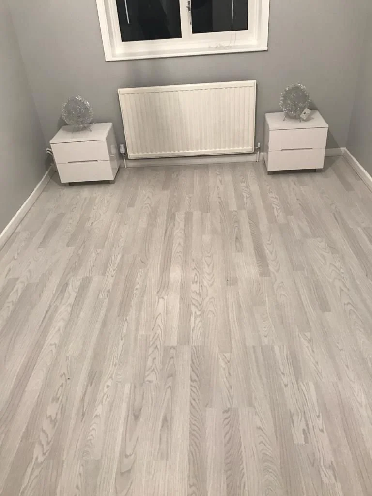 Empty room with light wood flooring, gray walls, a white radiator, two white side tables with decorative objects, and a window.