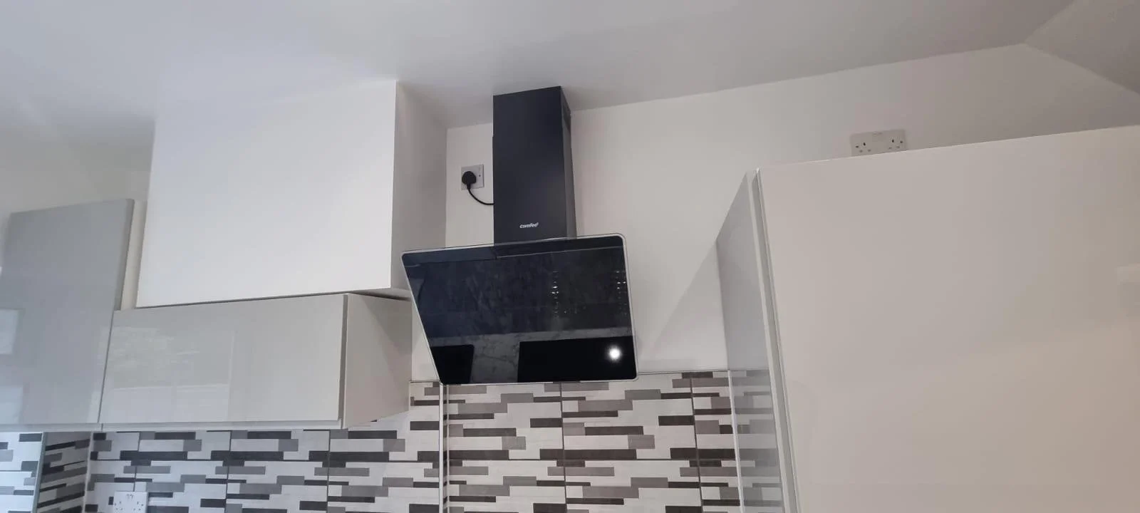 A kitchen with white upper cabinets, a black range hood, black stove, and a tiled wall with a gray, white, and black mosaic pattern.