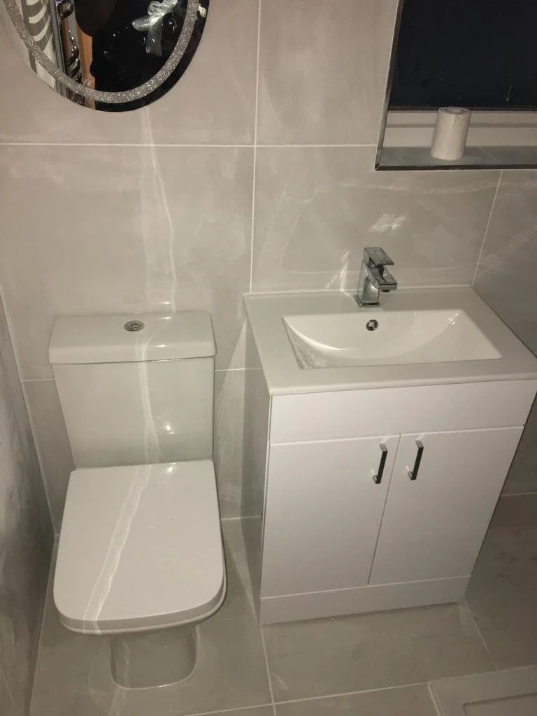 Small bathroom with a white toilet and a white vanity with a sink. There is a round mirror above the toilet and a small shelf with a roll of toilet paper.