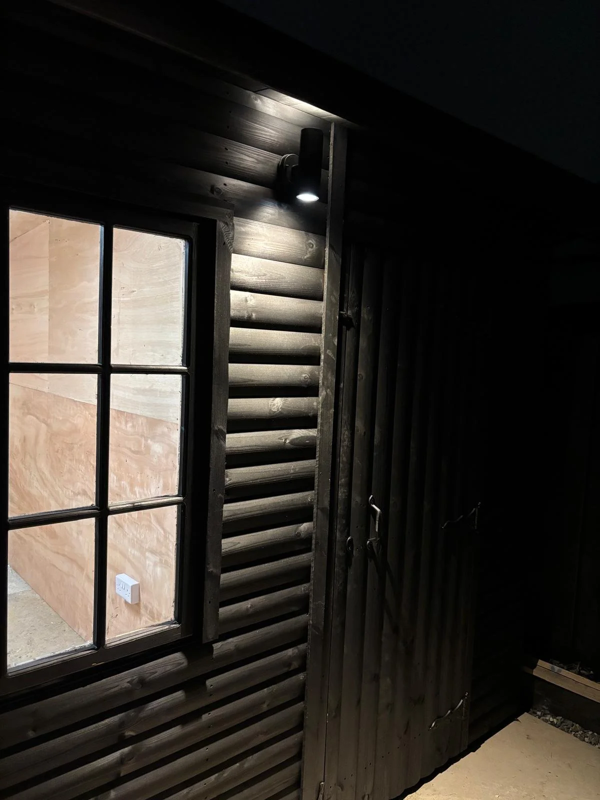 Close-up photo of a wooden cabin wall with a window and a door, illuminated by a small exterior light.