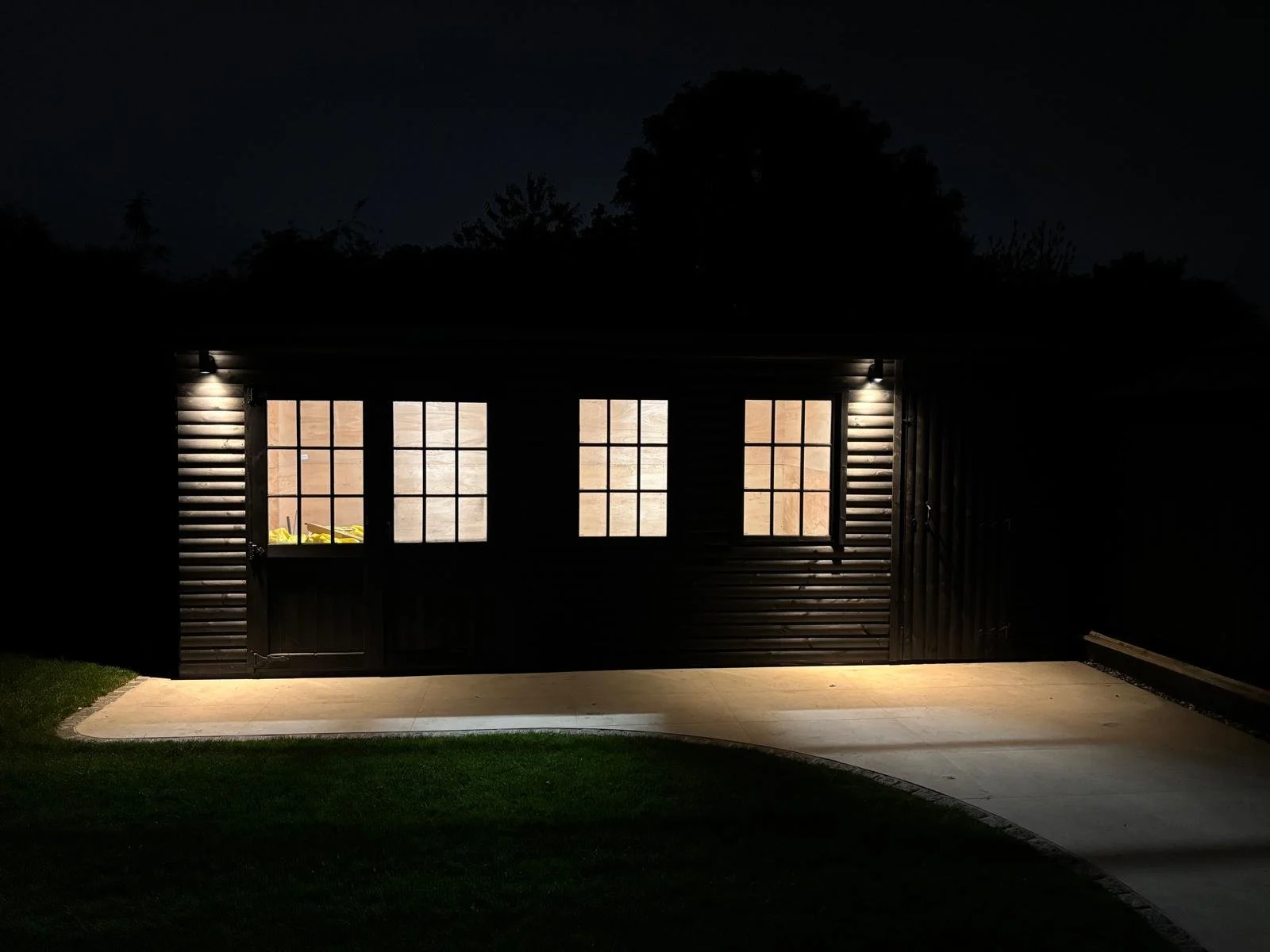 A garage with lit windows at night, showing some yellow objects inside. The exterior is black with horizontal siding, and the garage is connected to a curved driveway and a grassy area.