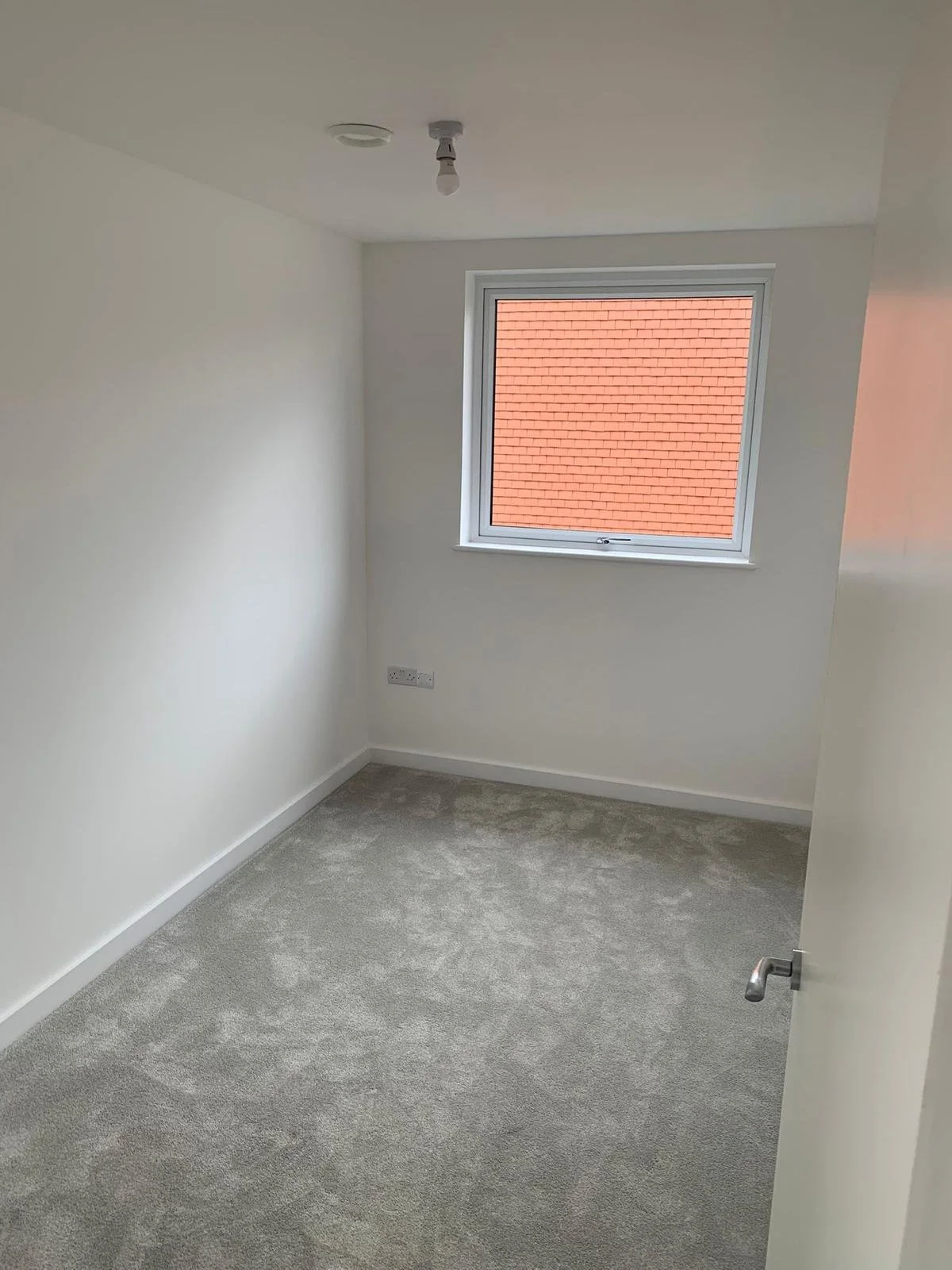 Empty room with white walls, gray carpet, a single window showing a red brick roof outside, and an exposed light socket on the ceiling.