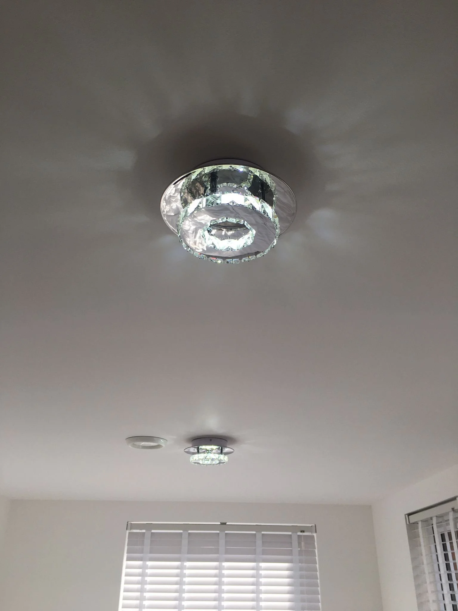 Ceiling with a modern crystal chandelier and two other ceiling fixtures, with a window having white blinds below.