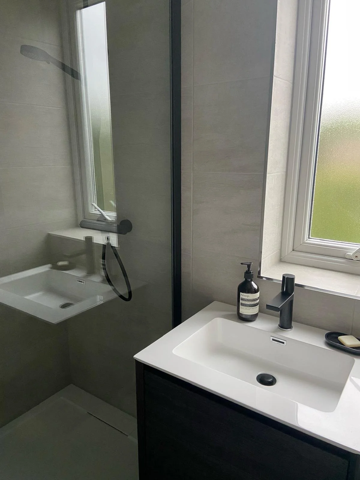 Modern bathroom with a sink, soap dispenser, and a window, mirror reflecting a shower area with a handheld showerhead.