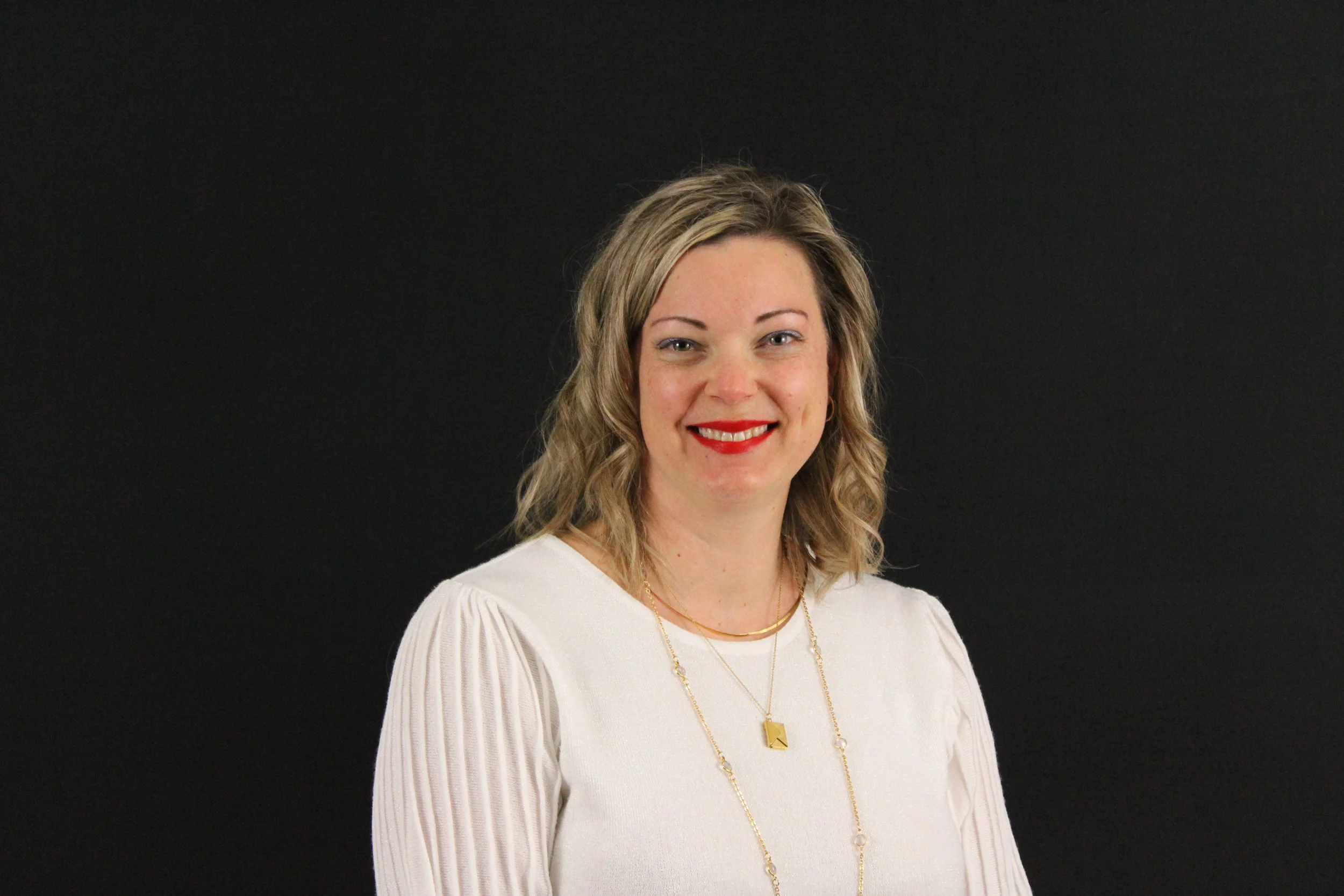 A woman with blonde curly hair smiling against a black background, wearing a patterned blouse and a gold necklace.
