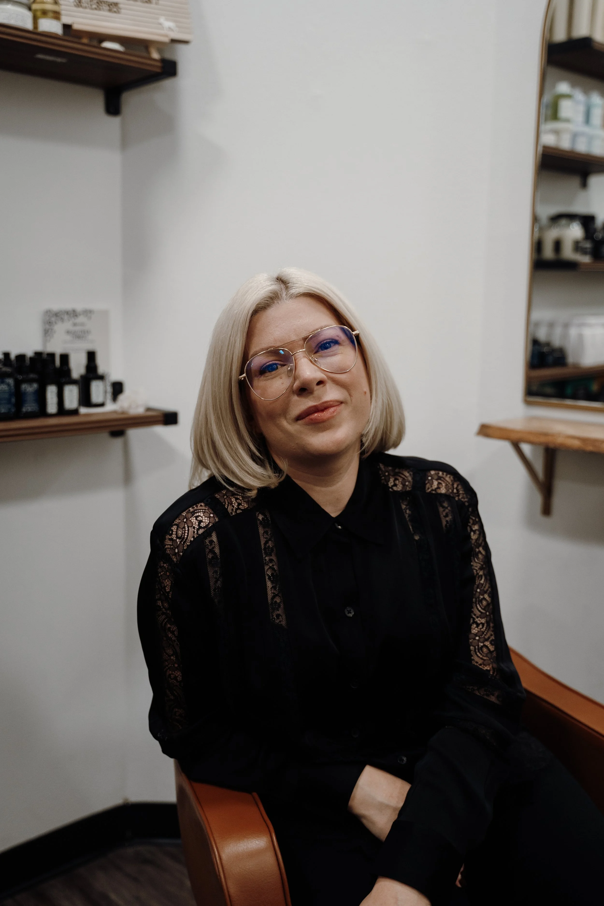 A young woman with long dark hair with red highlights, winking, sitting indoors against a textured beige wall, wearing a black denim jacket, a black shirt, and a smartwatch.