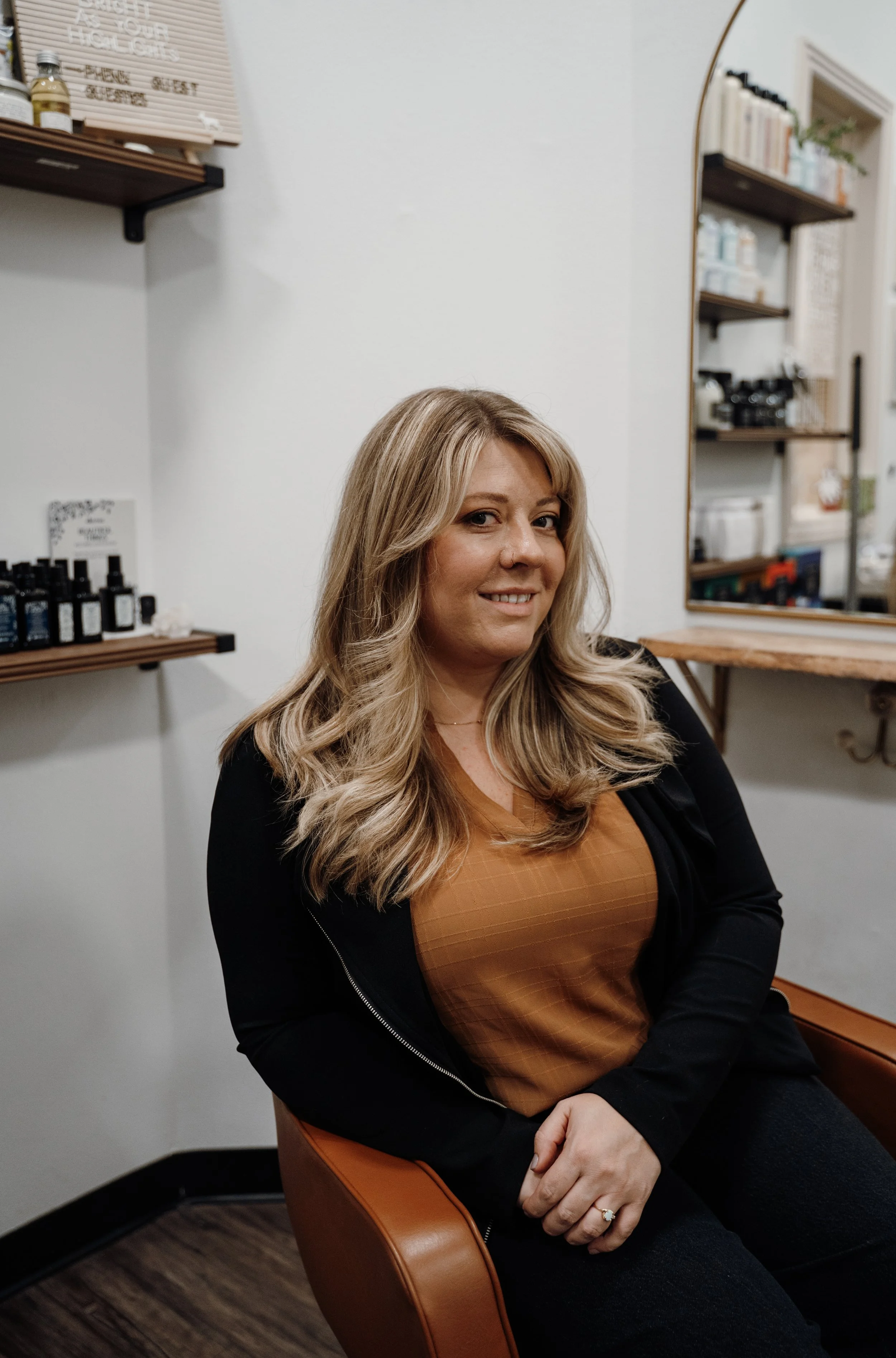A woman with long, wavy blonde hair seated in a salon, smiling at the camera, wearing a brown top and black jacket, with shelves of hair products and a mirror behind her.