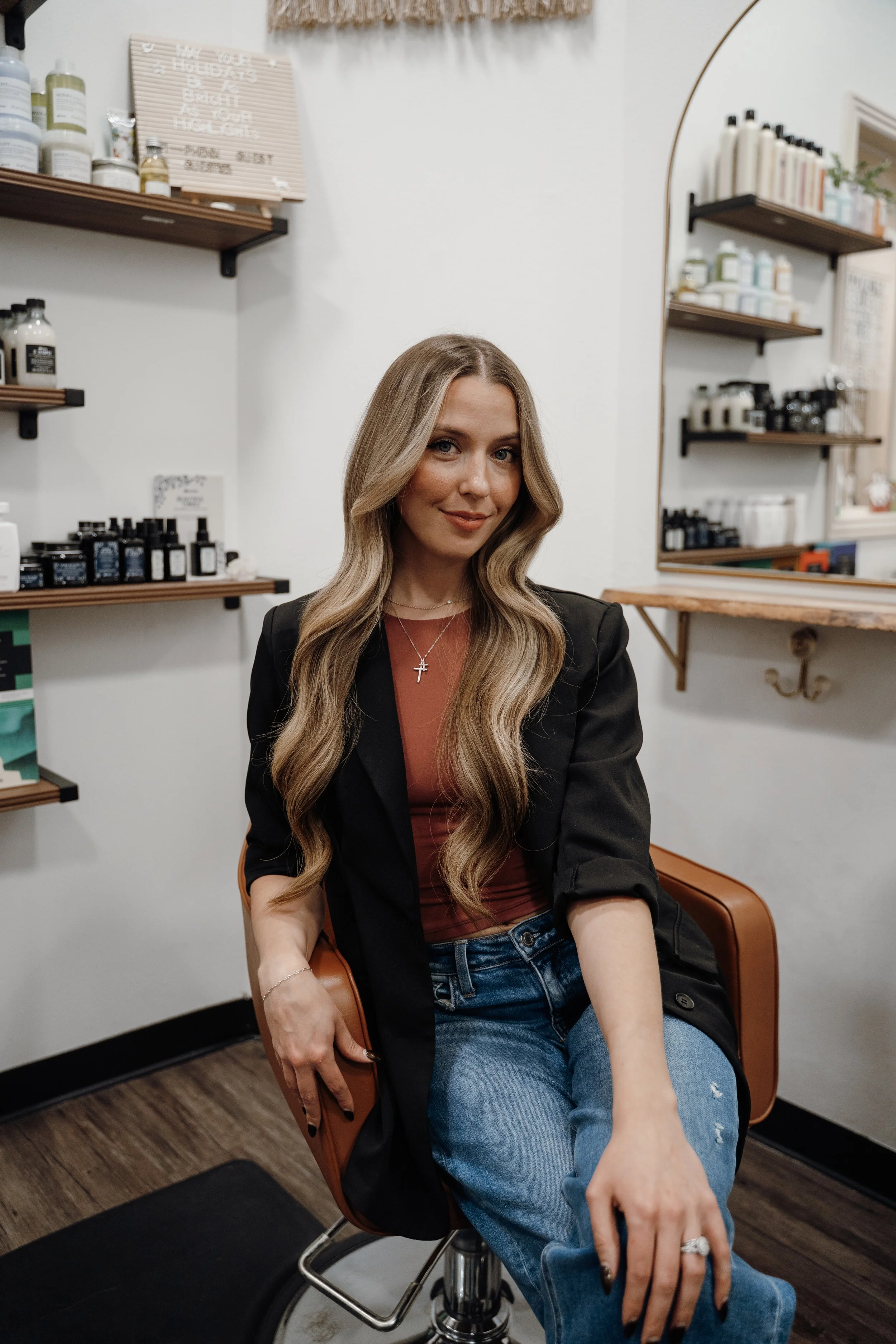 A woman with long wavy blonde hair and dark lipstick sitting on a wooden chair against a white wall, wearing a white t-shirt and ripped jeans.