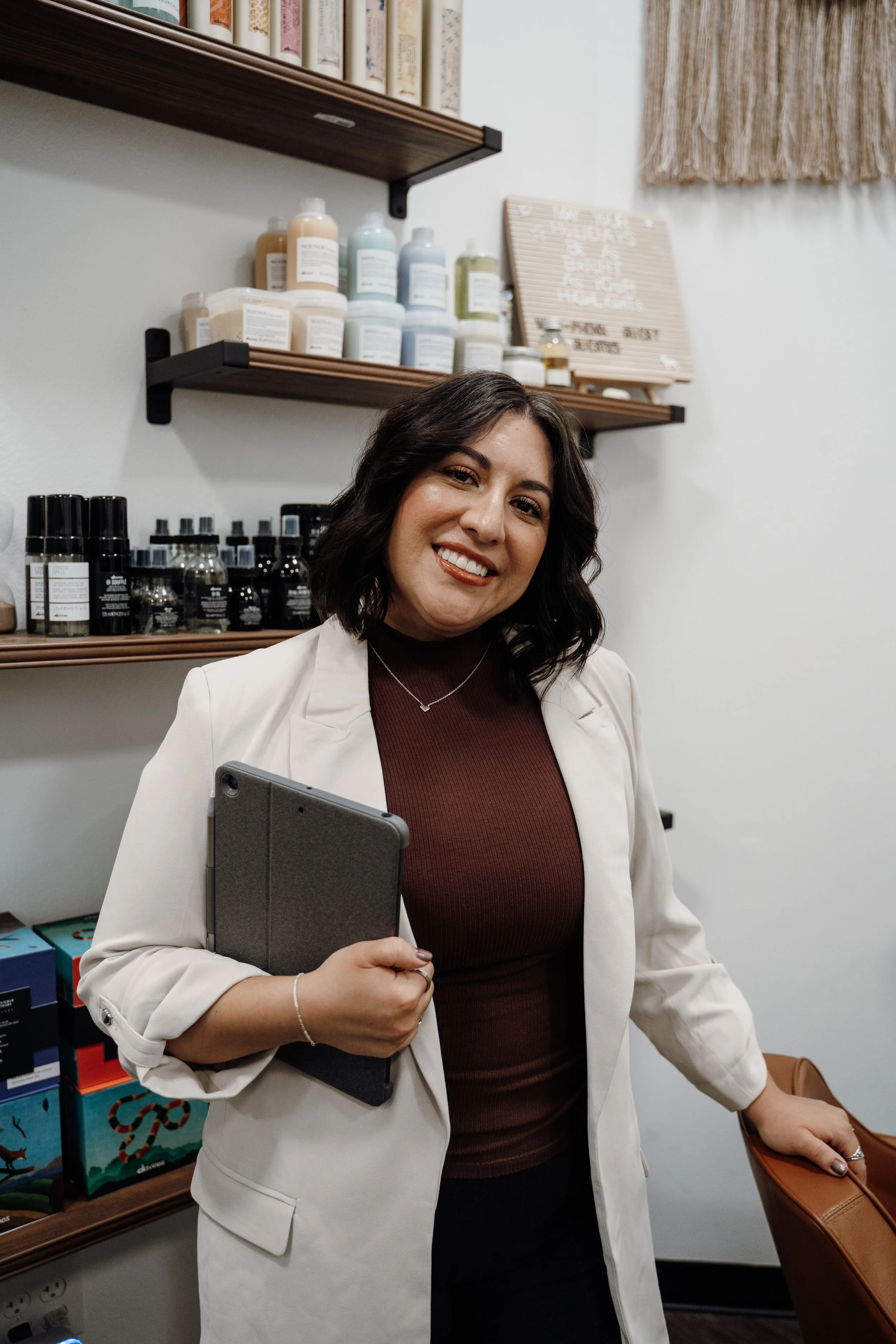 A woman with long, dark wavy hair smiling at the camera against a plain white background, wearing a beige tank top and ripped blue jeans.