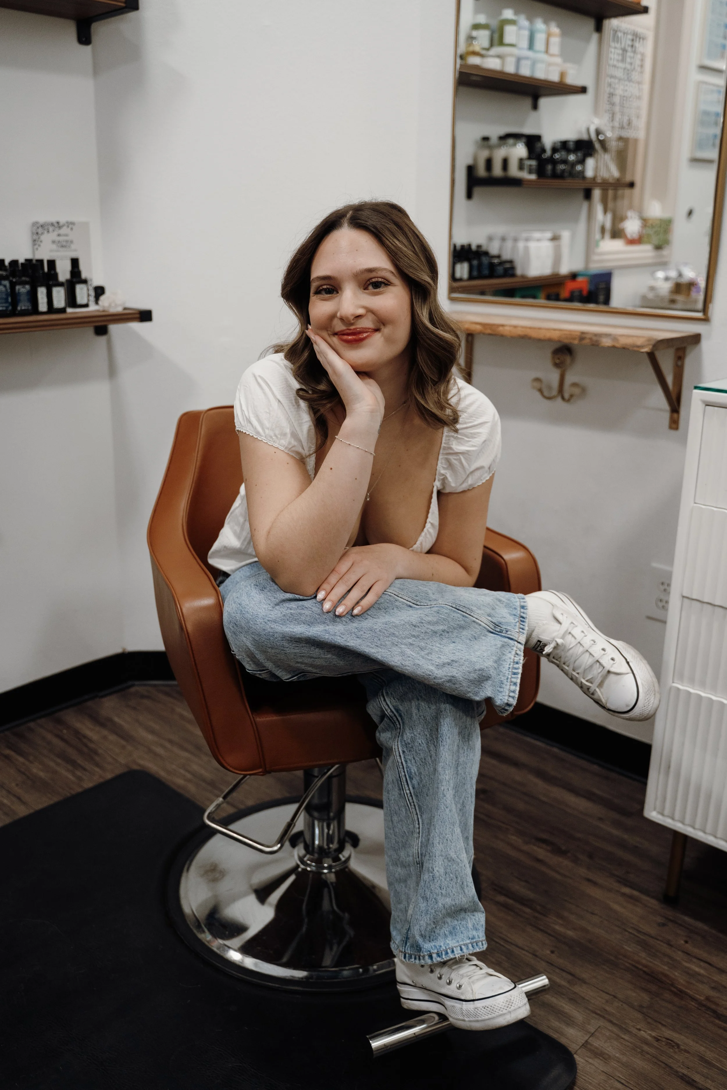Woman standing barefoot with hands on hips, wearing red glasses, white t-shirt, and blue jeans, in front of a white wall with an arch detail.
