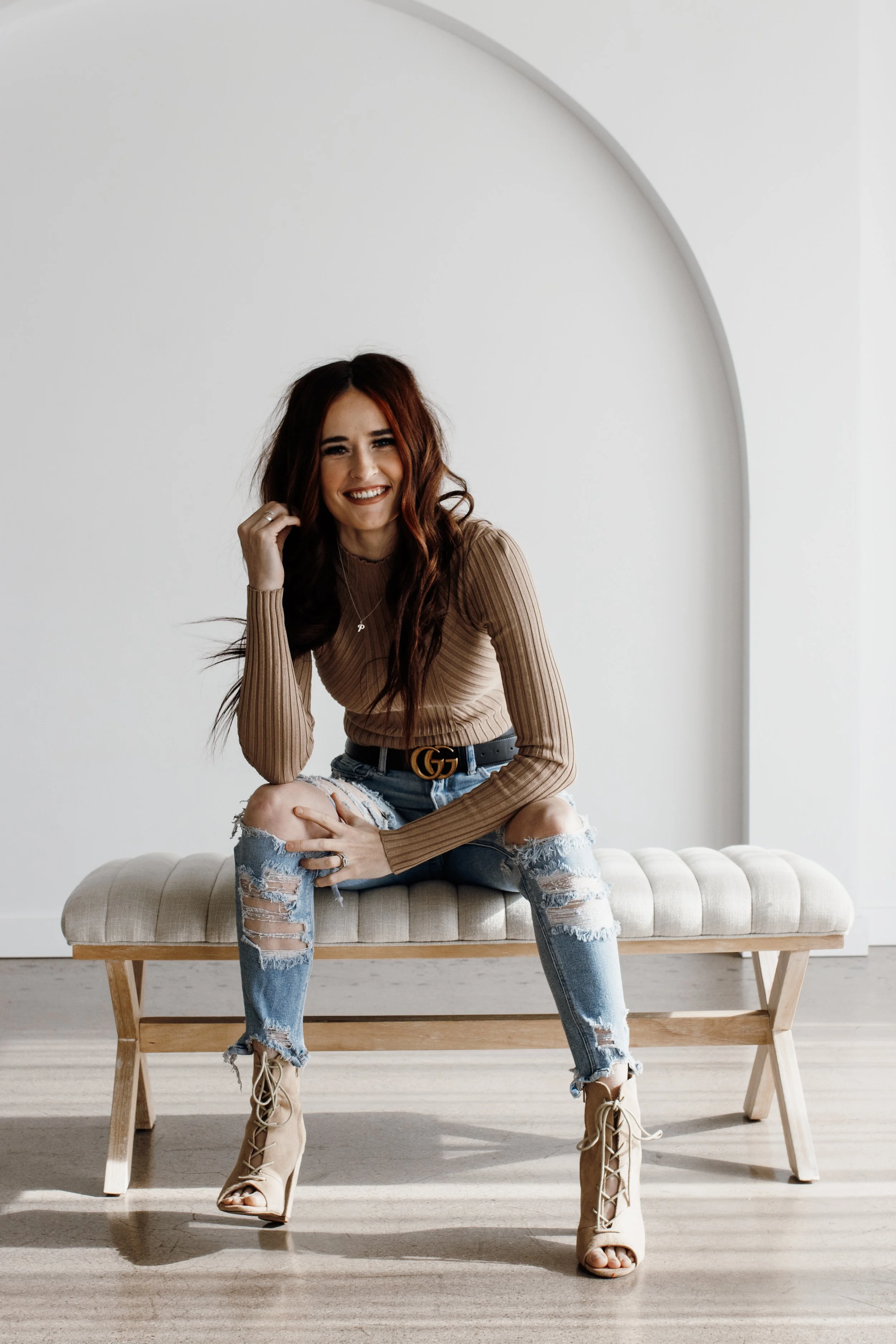 A woman with shoulder-length brown hair smiling, wearing a black long-sleeve top, leopard print skirt, and a necklace, standing in front of a beige chevron-patterned wall.