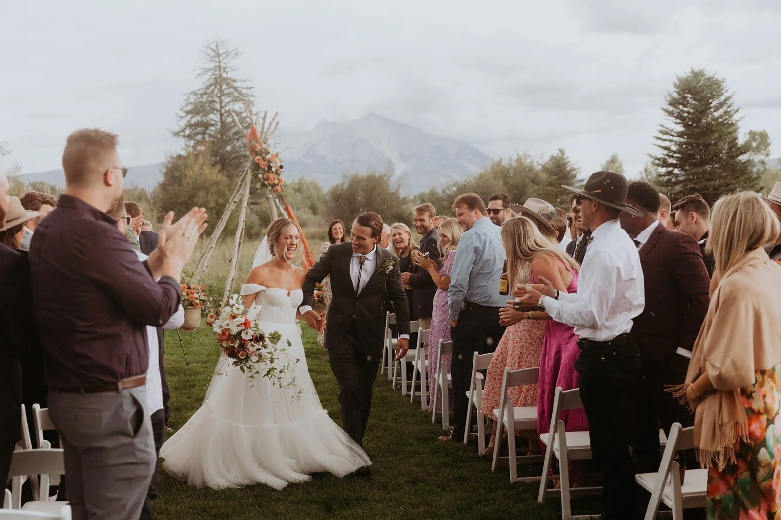 A bride and groom walking hand in hand down the aisle at their outdoor wedding ceremony, surrounded by guests clapping and smiling, with a scenic mountain and trees in the background.
