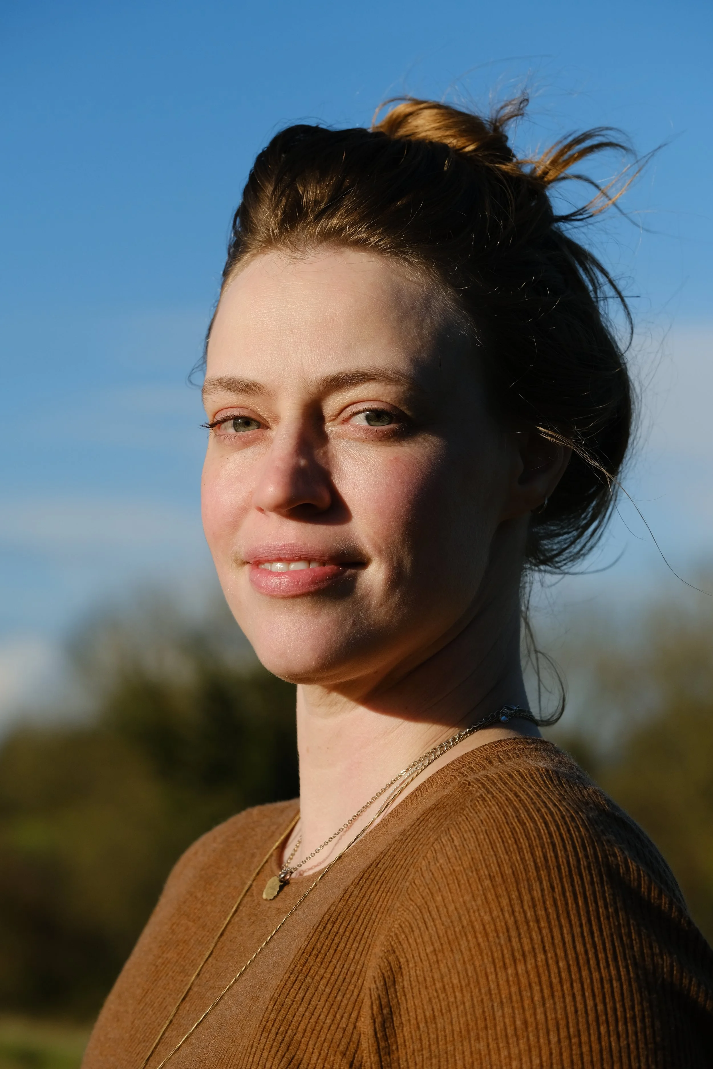 A woman with light skin and brown hair styled in a loose bun, smiling outdoors in sunlight, wearing a brown top and layered necklaces, against a backdrop of blue sky and trees.