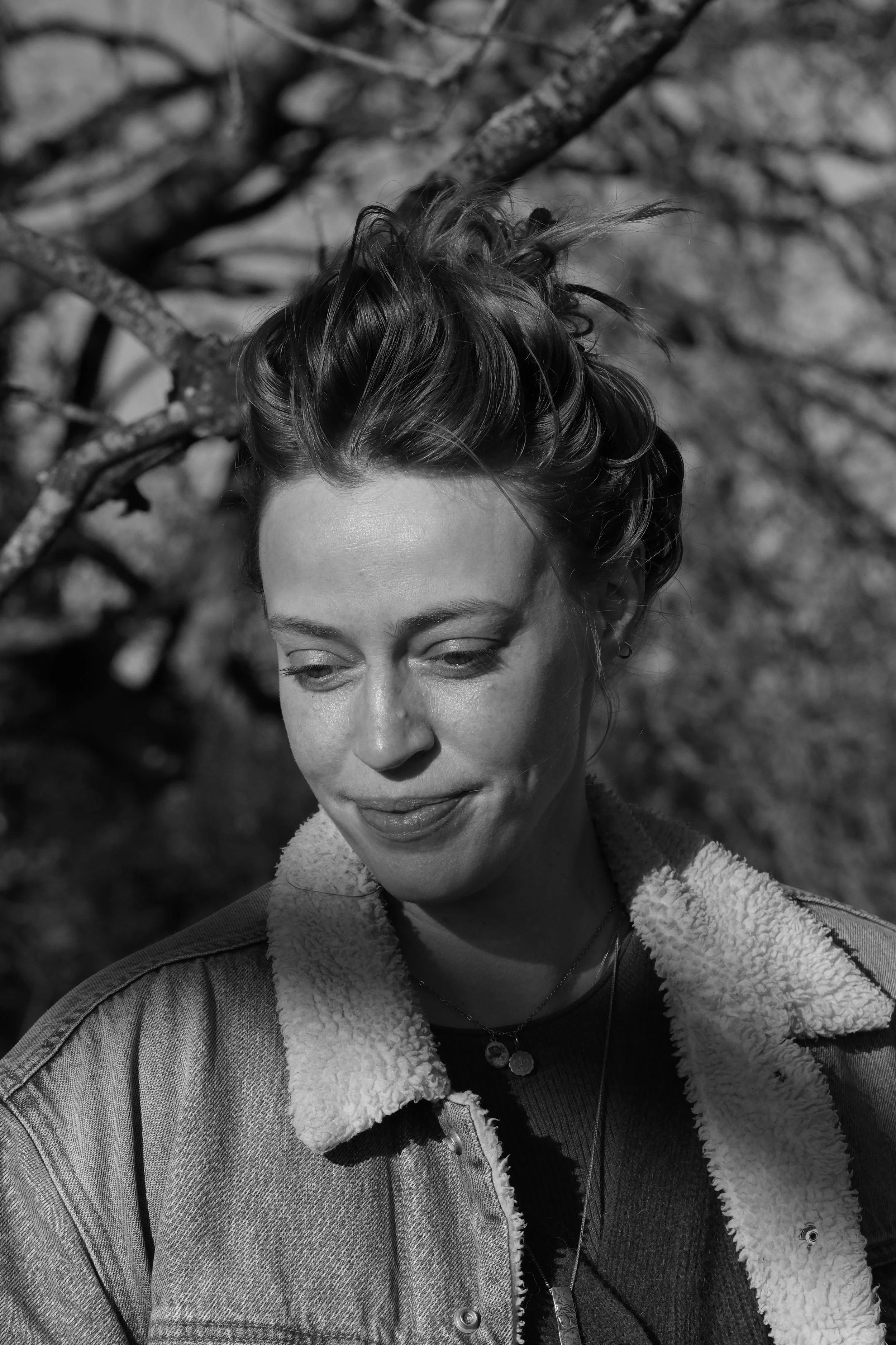 Black and white portrait of a woman with short wavy hair, smiling and looking downward, outdoors with tree branches in the background, wearing a denim jacket with a shearling collar and layered necklaces.