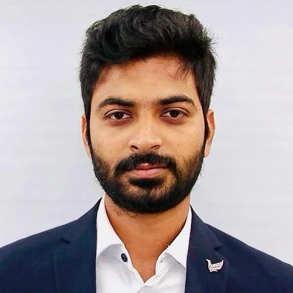 A young man with dark hair and a beard, wearing a navy blue suit jacket and a white shirt, standing against a light-colored background.