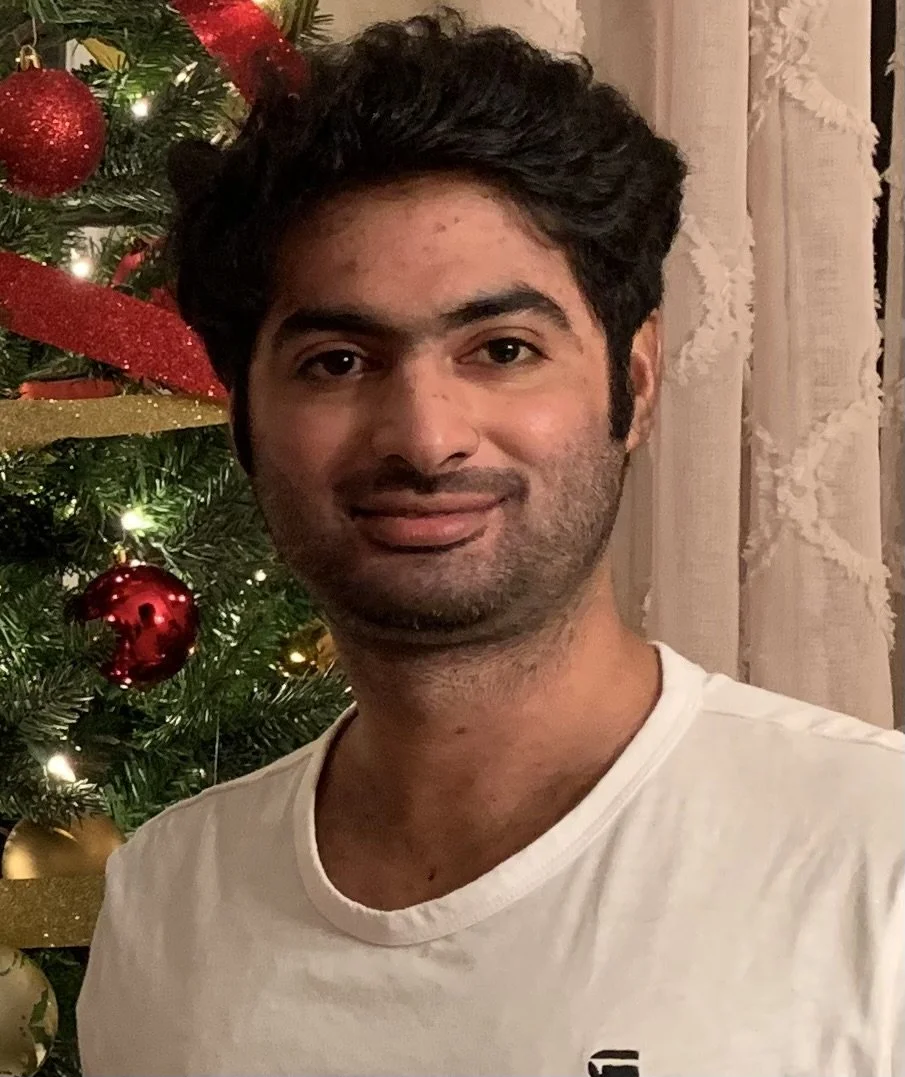 A smiling man with dark wavy hair standing in front of a decorated Christmas tree, wearing a white T-shirt.