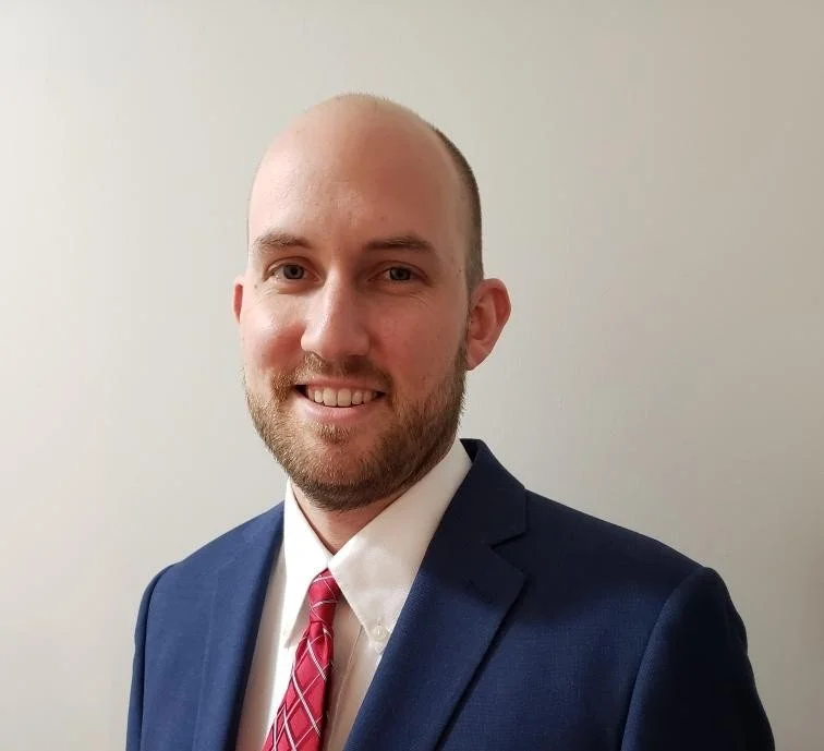 A smiling man with a beard, wearing a navy suit, white shirt, and red patterned tie, against a plain light-colored wall.