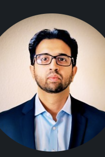 Professional portrait of a man with dark hair, beard, glasses, wearing a navy suit and light blue shirt, against a beige background.
