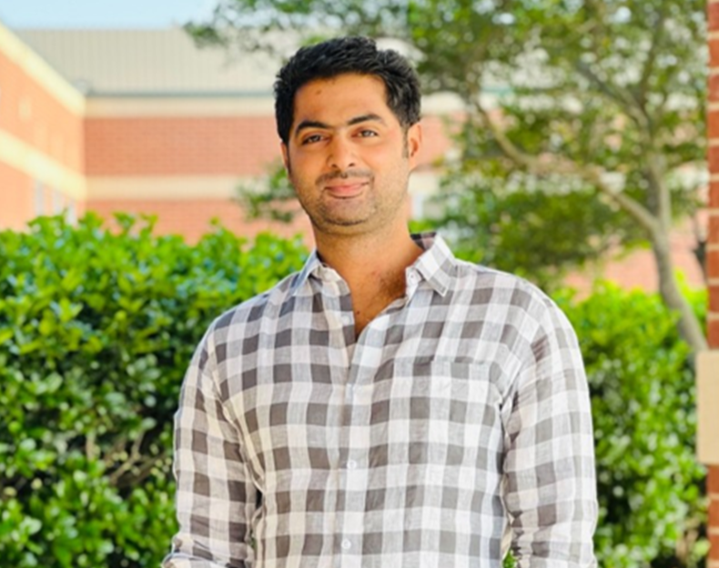 A smiling man with dark wavy hair standing in front of a decorated Christmas tree, wearing a white T-shirt.